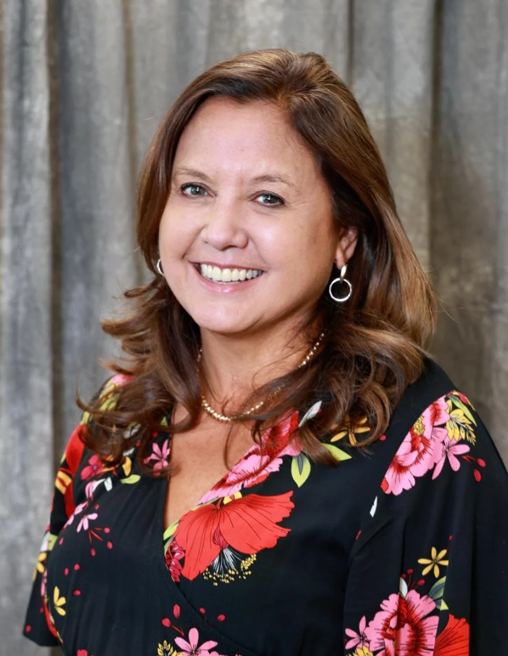Ramona Evey, a smiling woman with brown hair, wearing hoop earrings and a black floral blouse. She stands against a gray curtain background, conveying a cheerful tone.