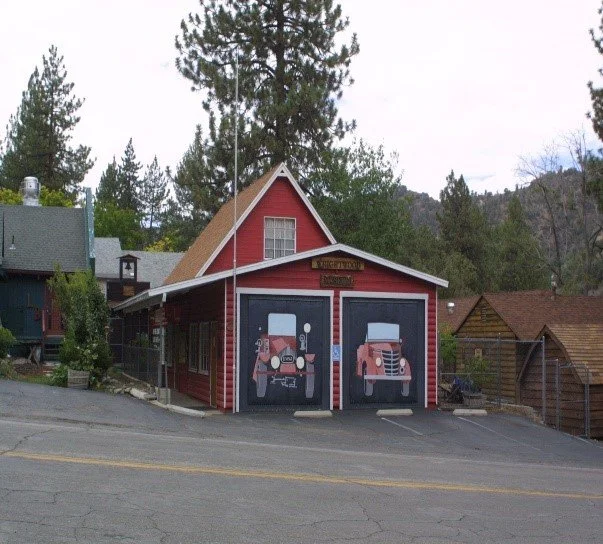 Wrightwood History Museum, a red building with vintage car murals on garage doors. Surrounded by trees, it conveys a nostalgic, quaint atmosphere, suggesting a small-town fire station.