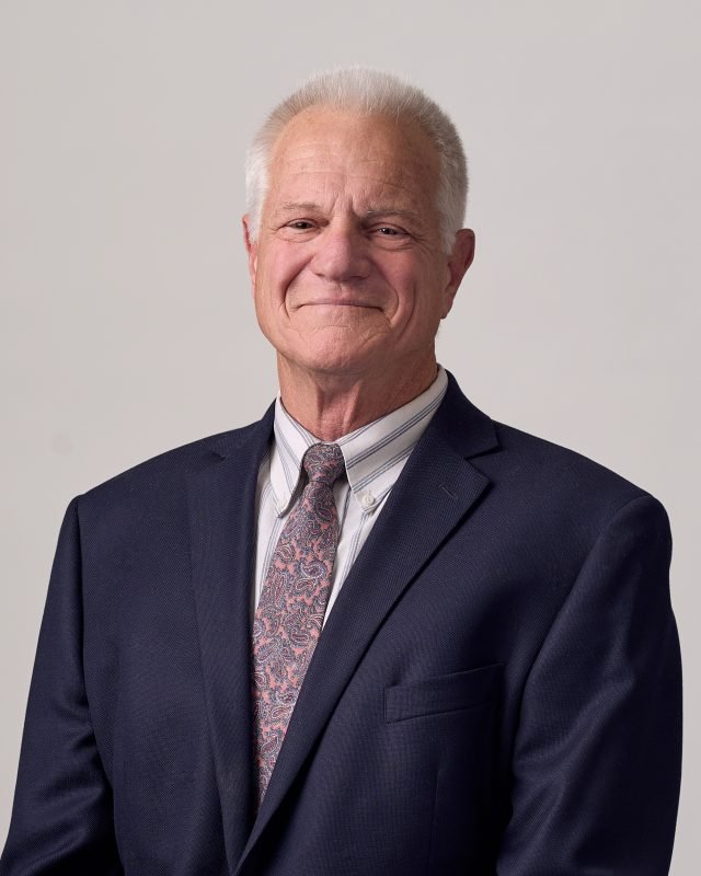 Rick Christensen, an elderly man with gray hair wearing a navy suit, striped shirt, and paisley tie, smiling warmly against a neutral background. Conveying professionalism.