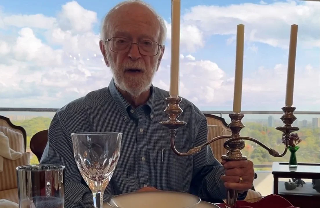 An elderly man with glasses and a beard sitting at a dining table holding a vintage candelabra with three candles, with wine glasses and a glass of water on the table, in a bright room with a large window and a cityscape view in the background.