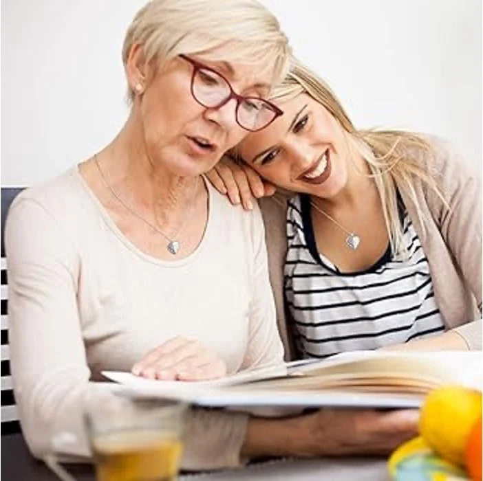 An elderly woman and a young woman are sitting together, looking at a book and smiling. The elderly woman has short gray hair, glasses, and is wearing a light-colored top. The young woman has long blonde hair, dark lipstick, and is wearing a striped shirt with a light cardigan. They seem to be enjoying a shared moment of reading and conversation.