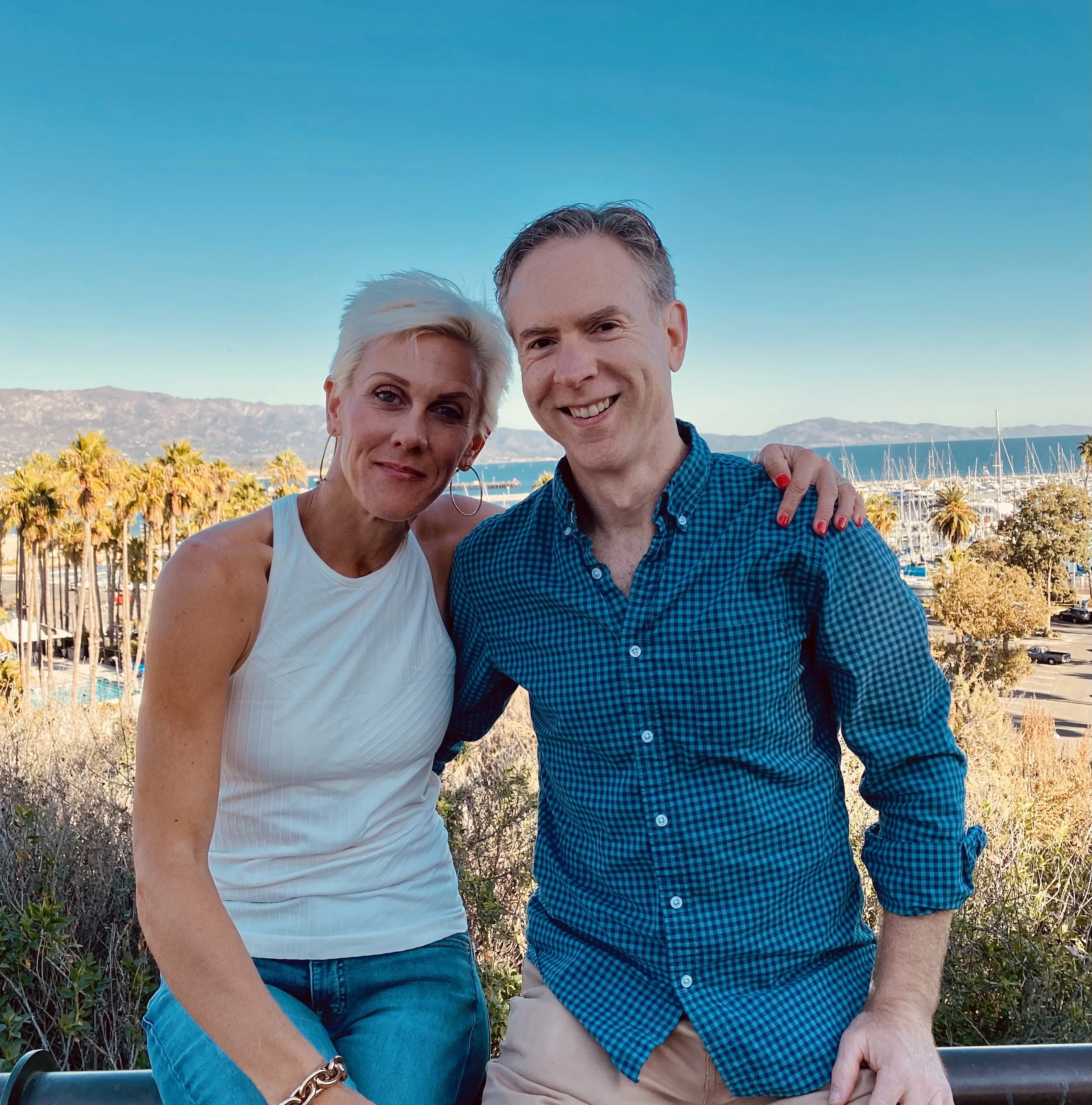 A woman with short blonde hair and a man with short gray hair smiling and posing together outdoors with a marina, sailboats, palm trees, and mountains in the background.