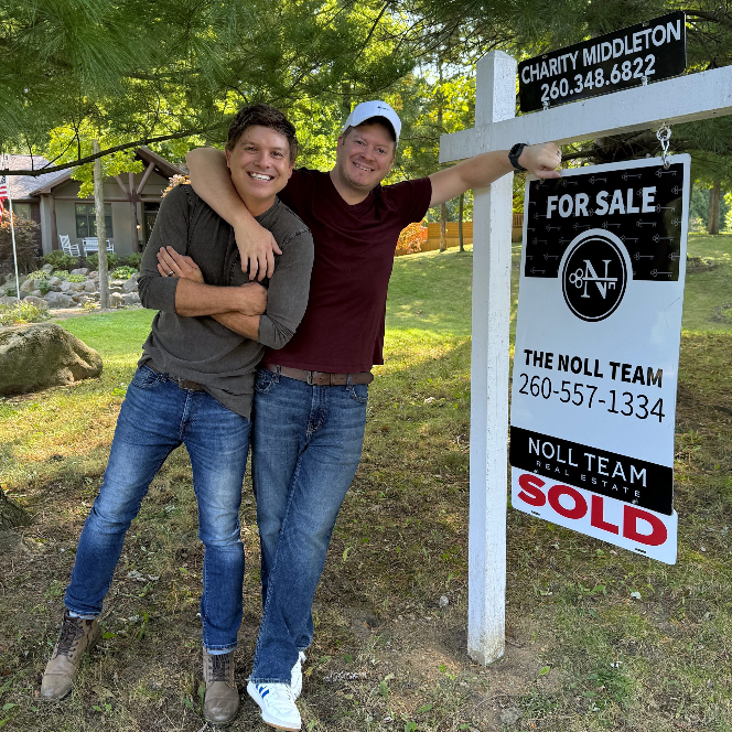 Two smiling men standing outdoors near a "For Sale" sign, with one arm around each other. One man in a gray shirt and jeans, the other in a maroon shirt and jeans, both wearing casual shoes.