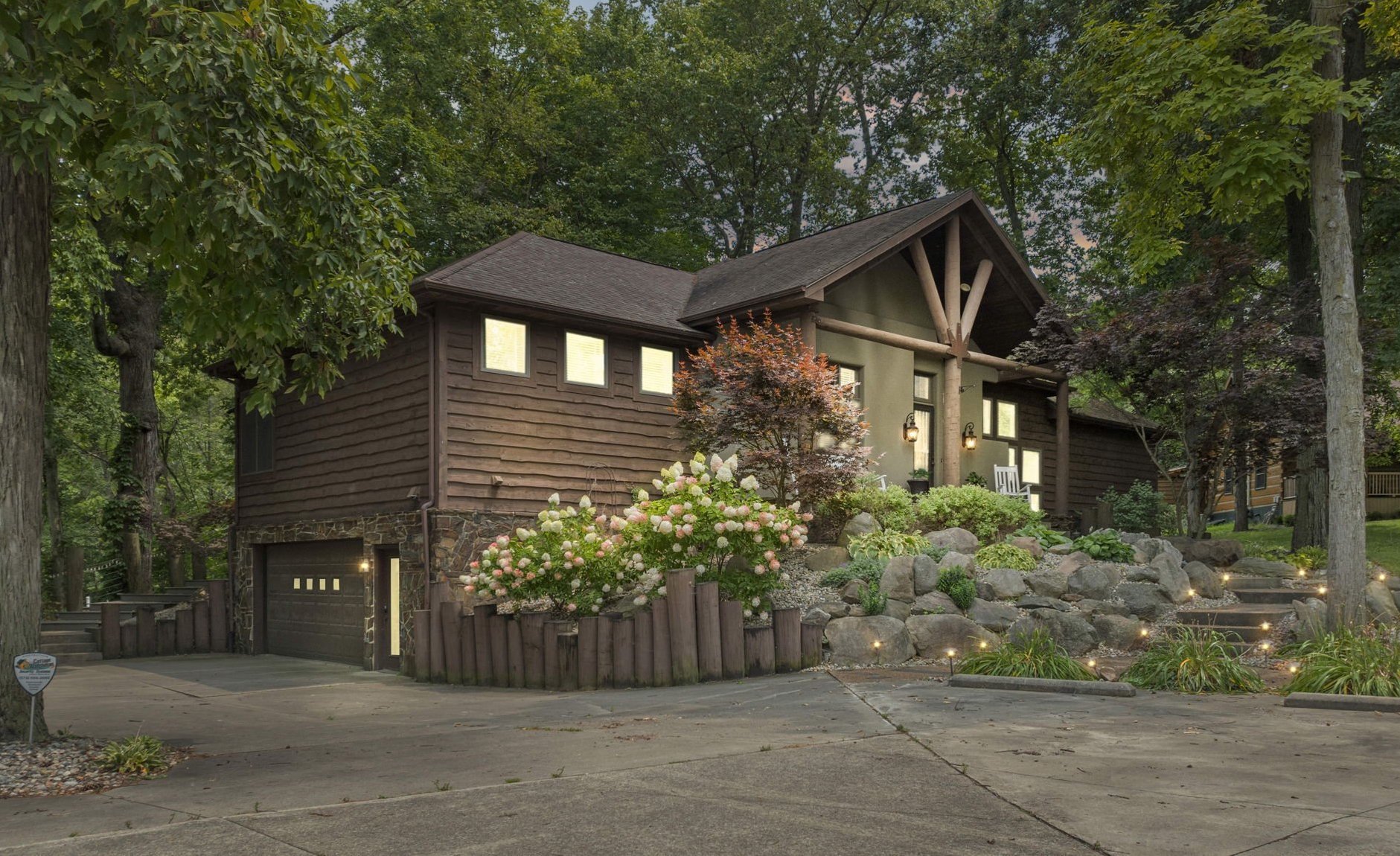 A house with a dark wood exterior and stone accents, surrounded by lush greenery and flowering bushes, with a driveway leading up to a garage, and outdoor lighting illuminating the landscape.