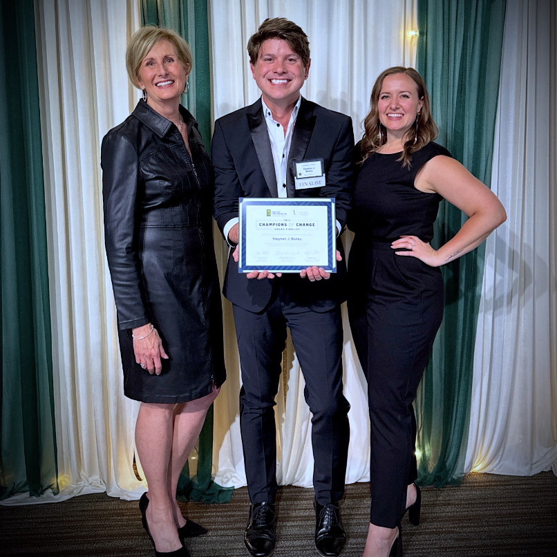 Three people stand together at an award ceremony, with a woman on the left, a man in the middle holding a certificate, and a woman on the right. The background features curtain drapes in cream and green.