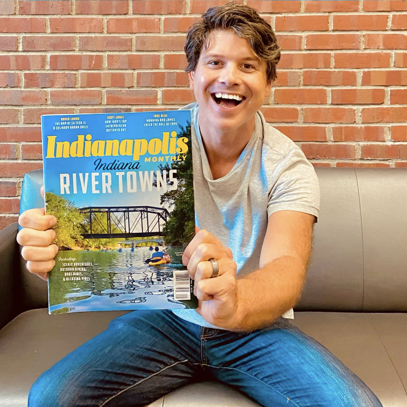 A smiling man holding a magazine titled 'Indianapolis Monthly' with a cover image of a river scene and a bridge, sitting on a gray couch against a brick wall.