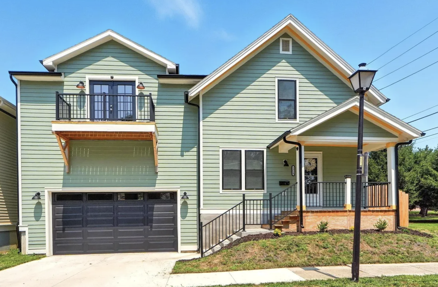 A two-story house with light green siding, black garage door, front porch, black railing, and a balcony with black railing and double doors, under a blue sky.