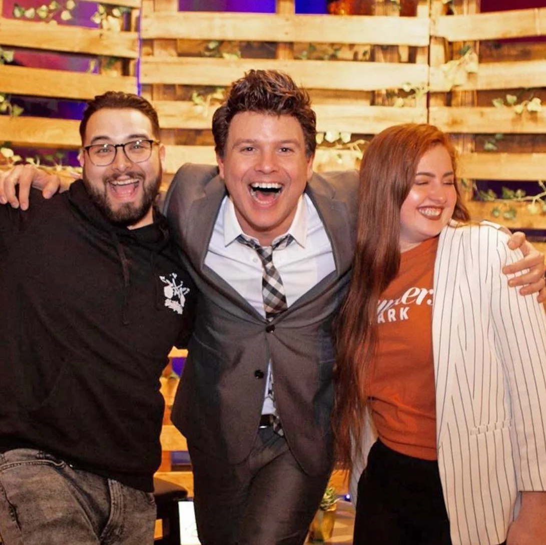 Three people smiling and posing together, with a wooden background and some greenery.