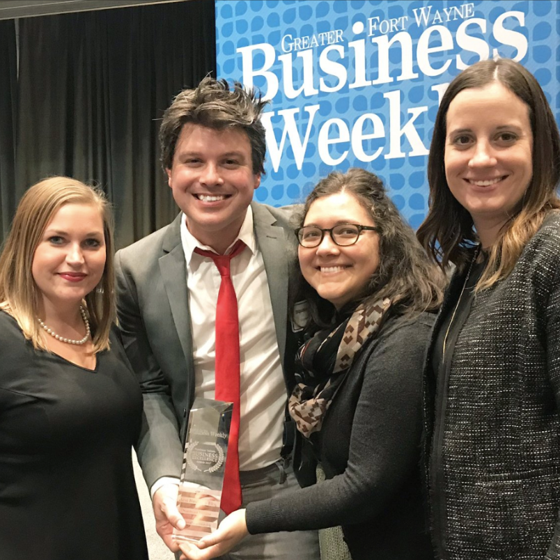 Four people smiling at a business event with a banner in the background that reads 'Greater Fort Wayne Business Week.' One person holds a trophy or award.