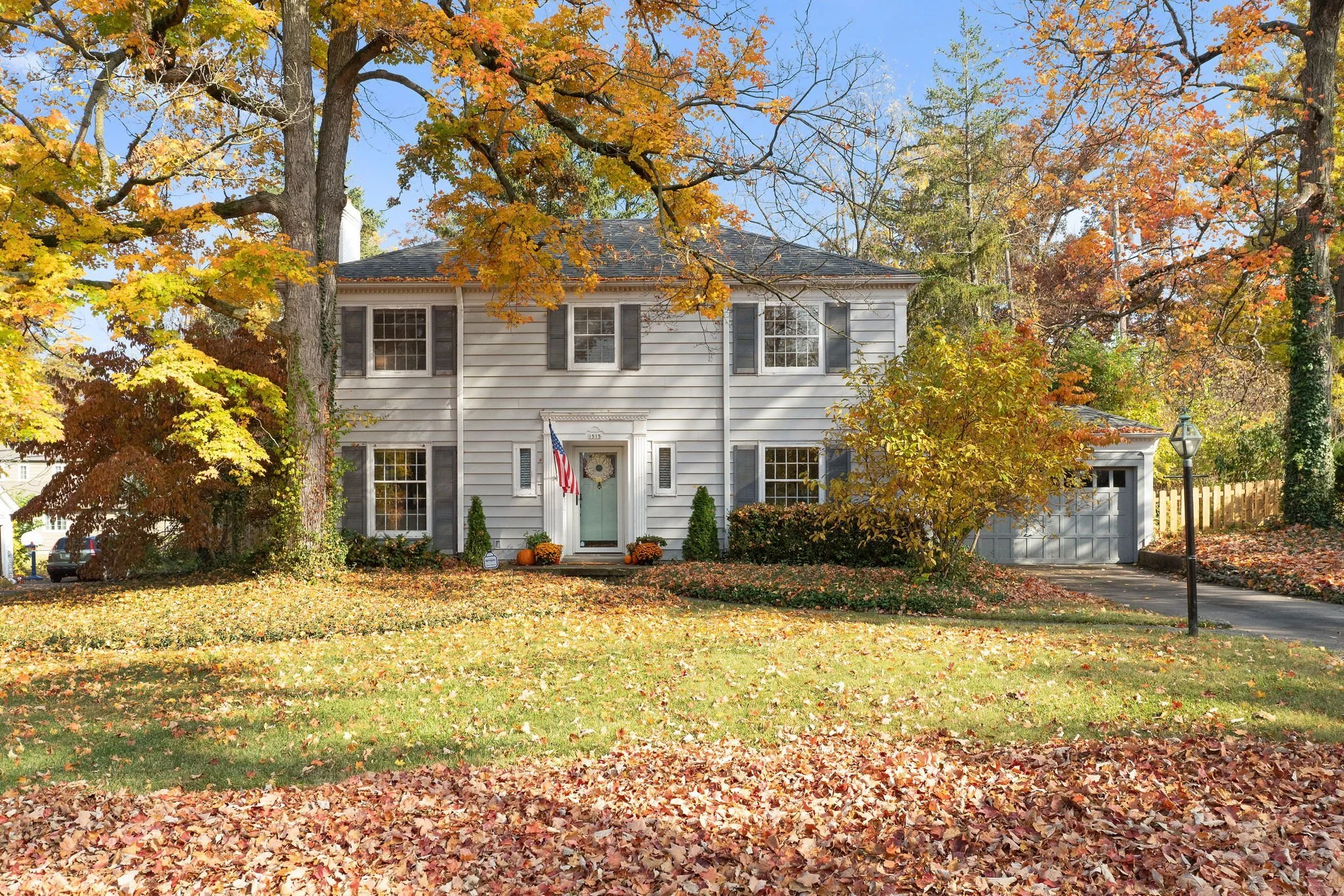 A white two-story house with gray shutters and a front door adorned with a wreath, surrounded by fall foliage with colorful trees and fallen leaves on the ground, and a driveway leading to a garage on the right.