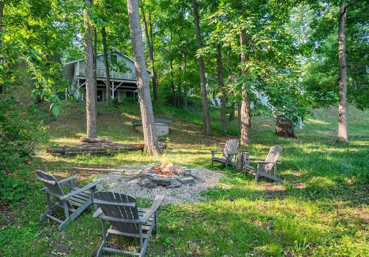 A backyard with a campfire ring surrounded by four wooden chairs on a grassy area, with trees and a house on stilts in the background.