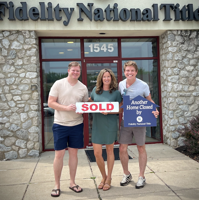 Three people standing in front of a real estate office, smiling and holding signs that say 'SOLD' and 'Another Home Closed by Fidelity National Title.'