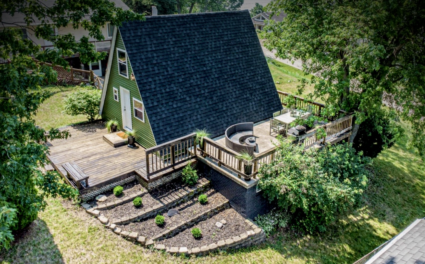 Aerial view of a backyard with a green tiny house, wooden deck, outdoor seating, and landscaped garden with plants and trees.