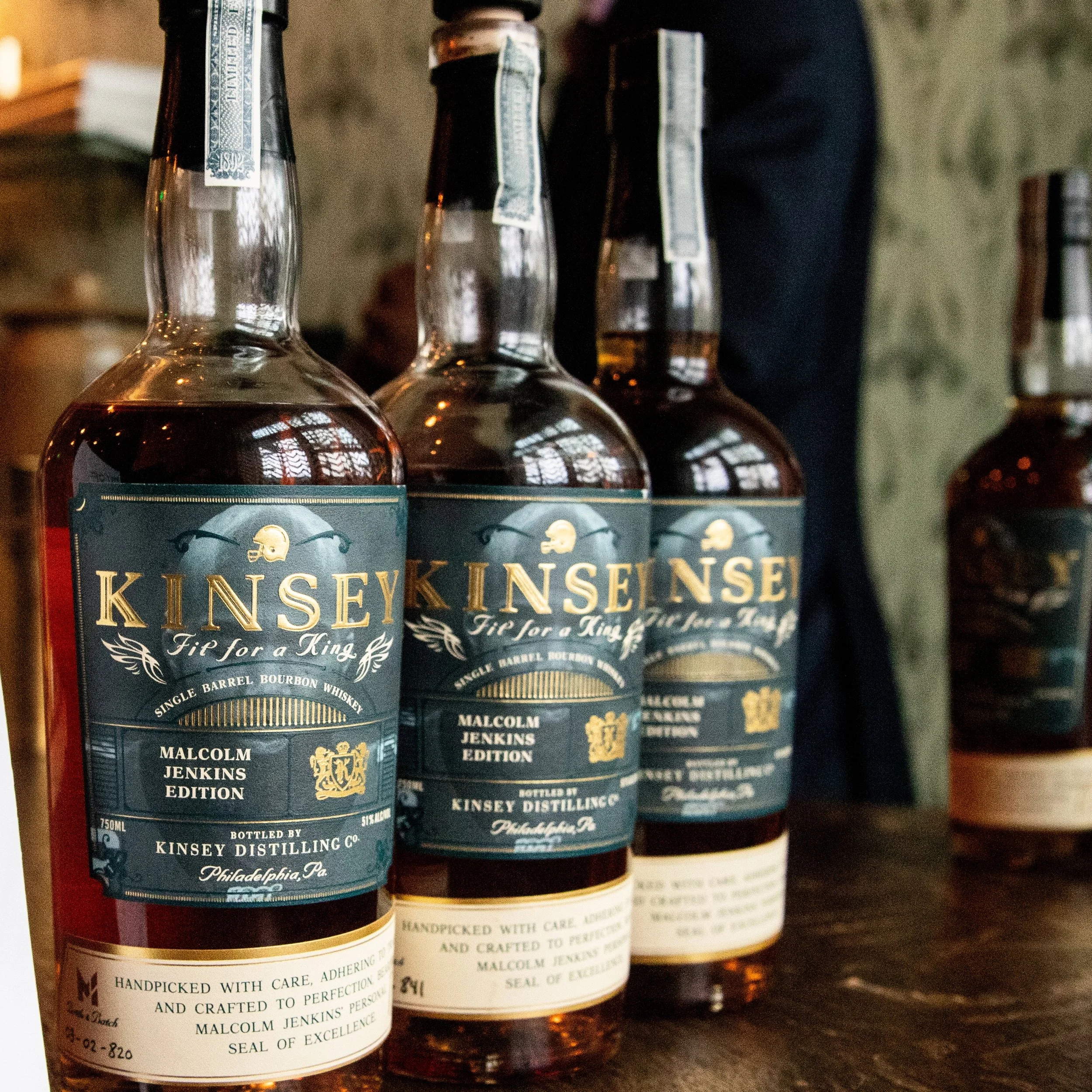 Three bottles of Kinsey tip for a king bourbon whiskey on a wooden surface with a person partially visible in the background.