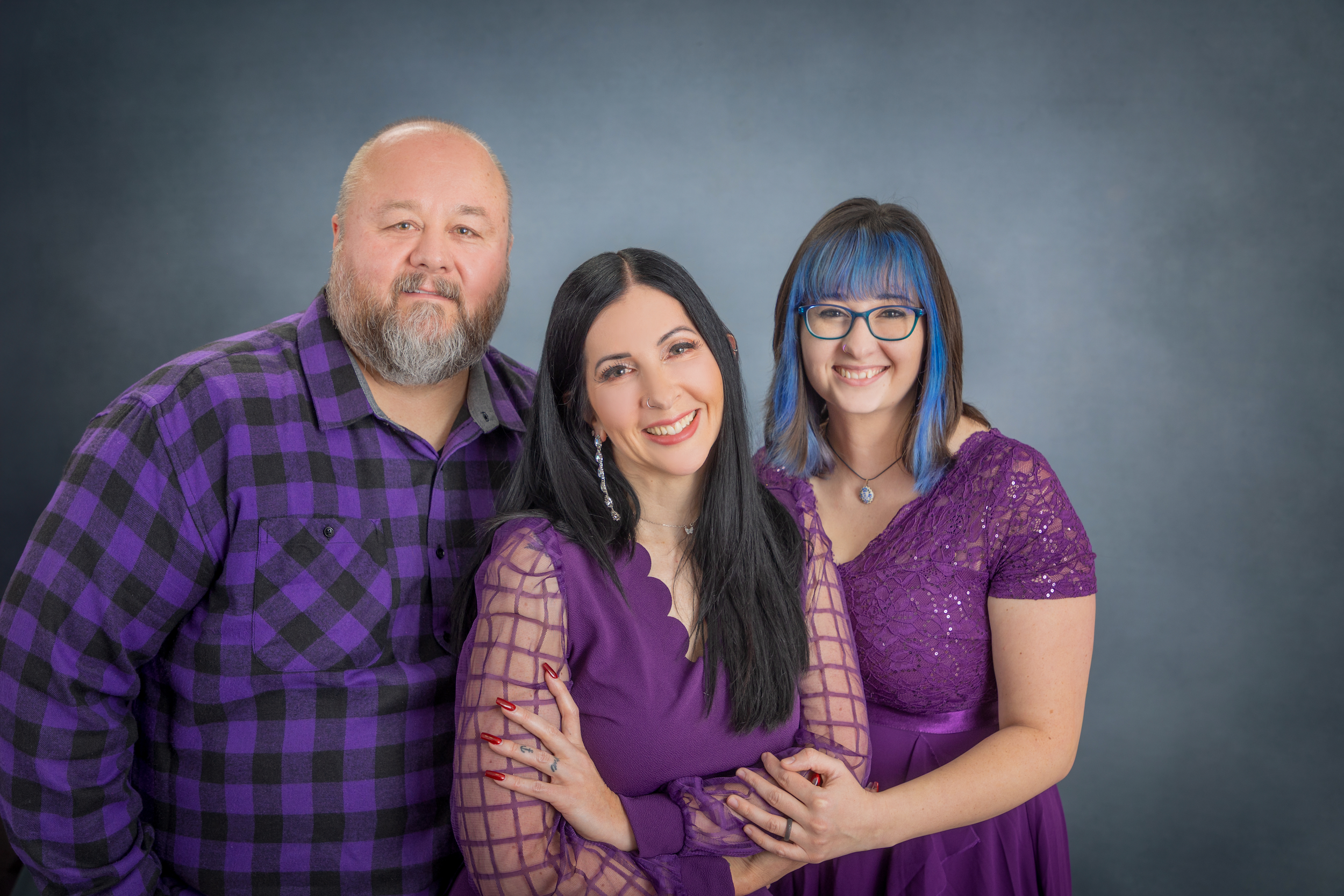 A group of three people, a man and two women, smiling and posing together against a dark gray background. All are dressed in purple clothing.