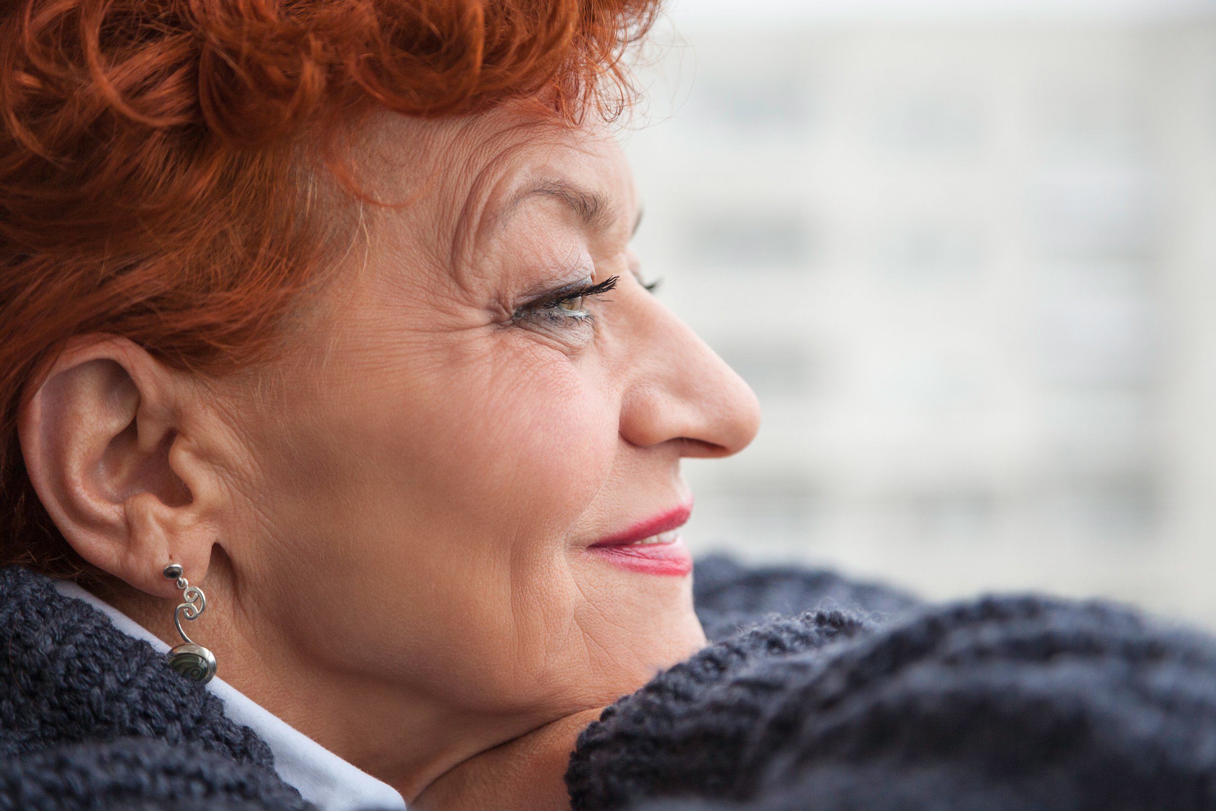 Close-up side profile of a woman with red hair, earrings, and a textured dark jacket, looking thoughtfully out a window with a blurred cityscape background.