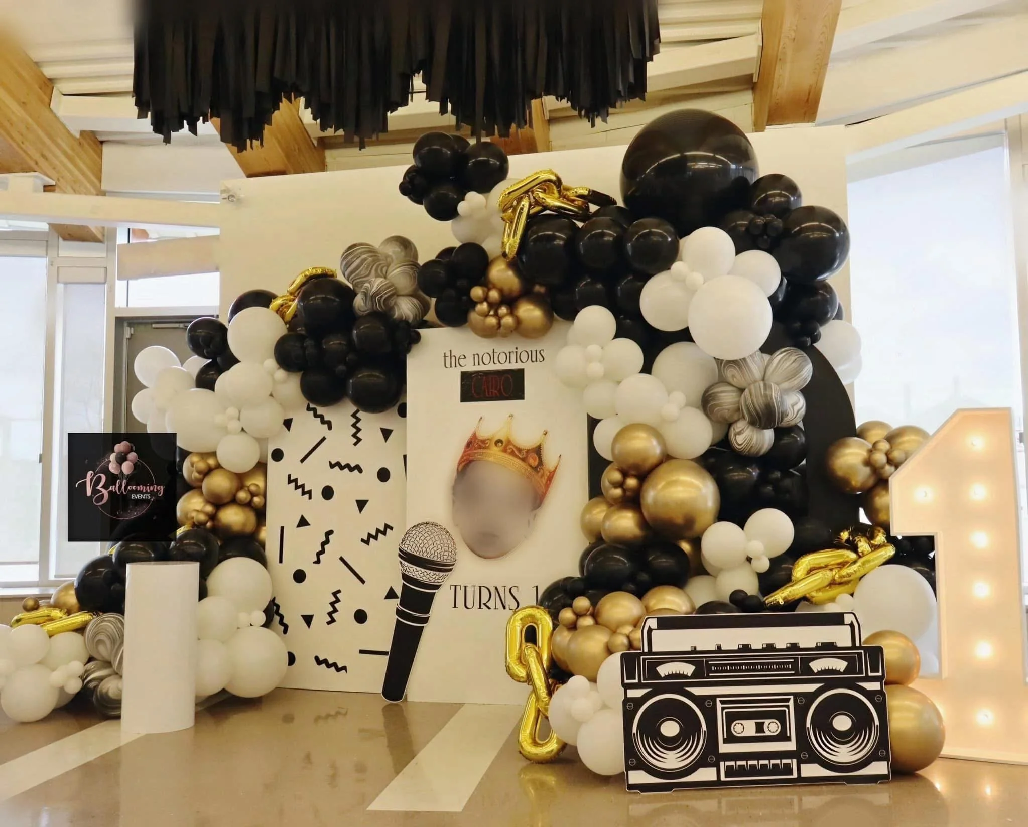 Balloon decoration for a 10th birthday celebration with black, white, gold, and gray balloons, a sign with a crown and microphone, a large illuminated number 10, a boombox cutout, and a poster with a face, crown, and the text 'the notorious CALRO TUR