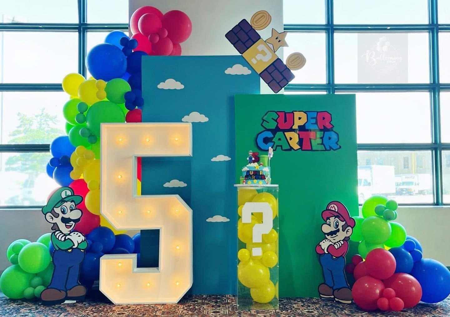 Decorations inspired by Super Mario Bros., featuring characters Mario and Luigi, with colorful balloons, large letter sign, clouds, question block, and a cake with Mario and Luigi toppers.