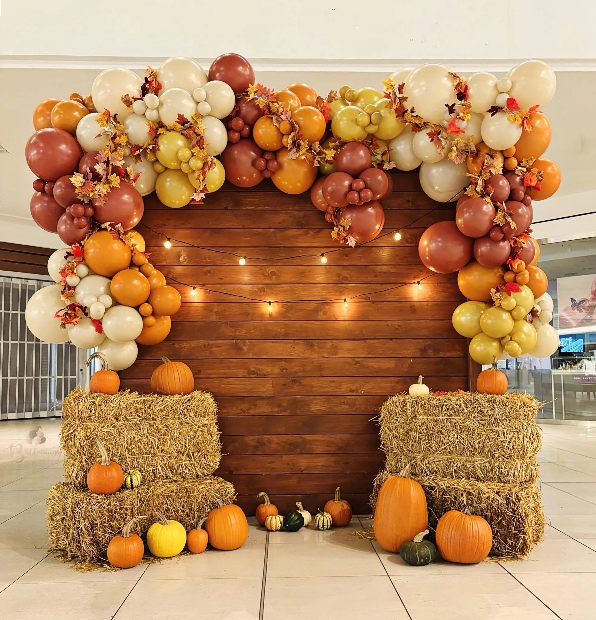 Fall-themed display with a wooden backdrop, containing a balloon garland in autumn colors, hay bales, and pumpkins of various sizes and colors, decorated with fall leaves and string lights.