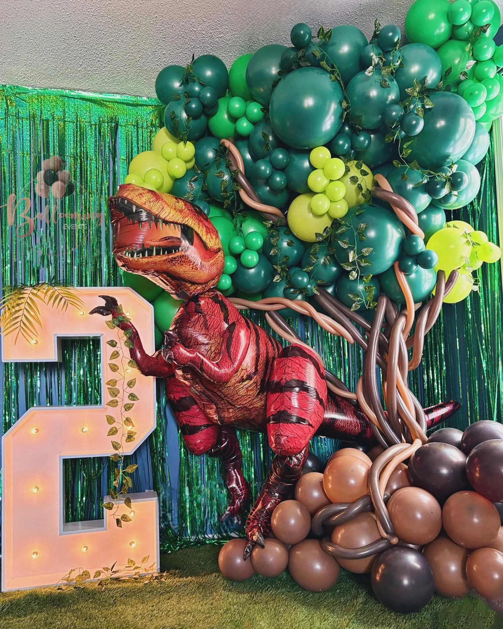 Decorative backdrop featuring a T-Rex dinosaur figure, green balloons, and a large illuminated number 2, with shiny green and brown strings of balloons and a green tinsel curtain.