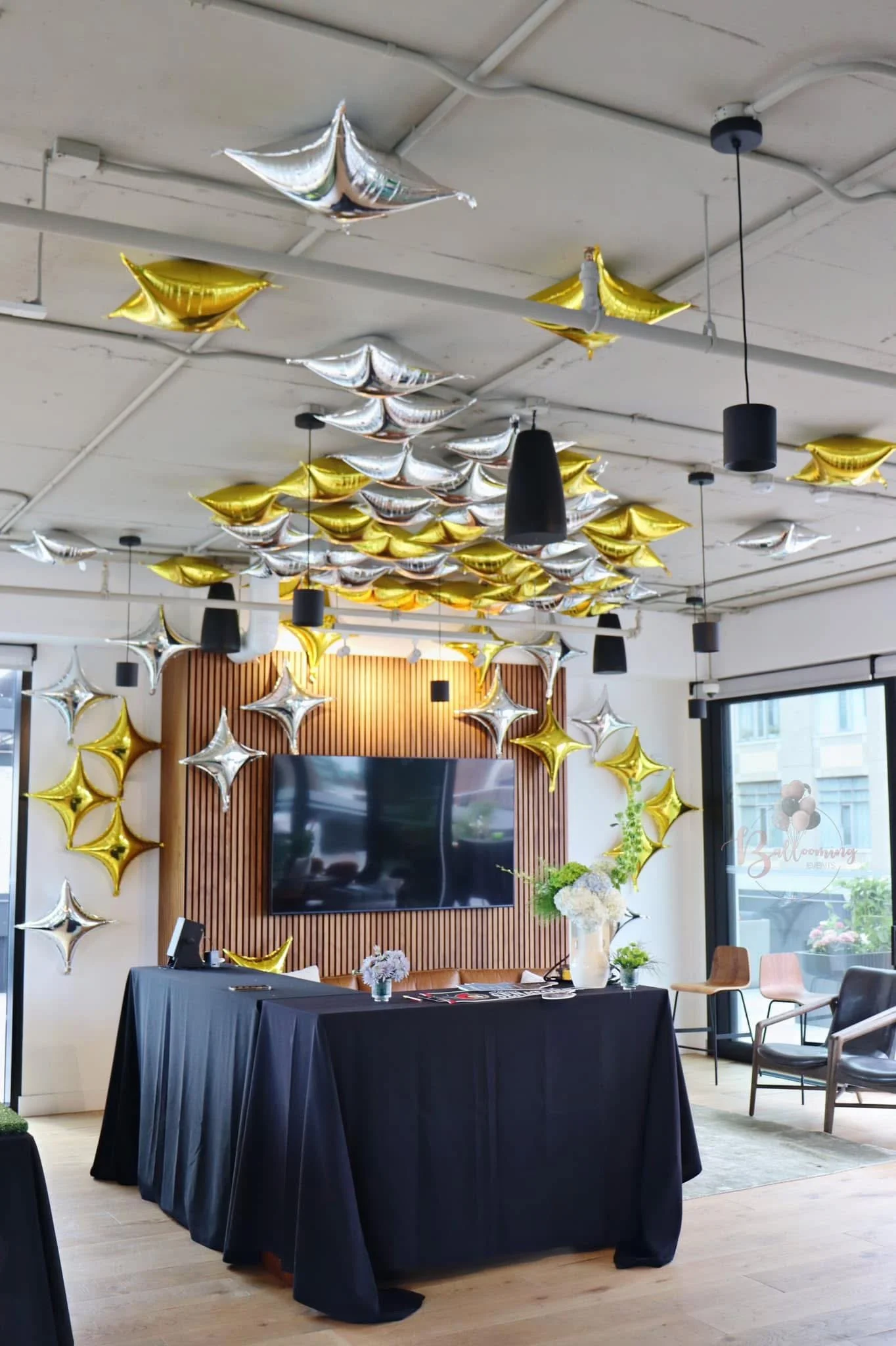 Interior decorated with gold and silver star-shaped balloons on the ceiling and walls, with a large flat-screen TV mounted on a wood-paneled wall and a table covered with a black cloth in front, adorned with flowers in a vase.