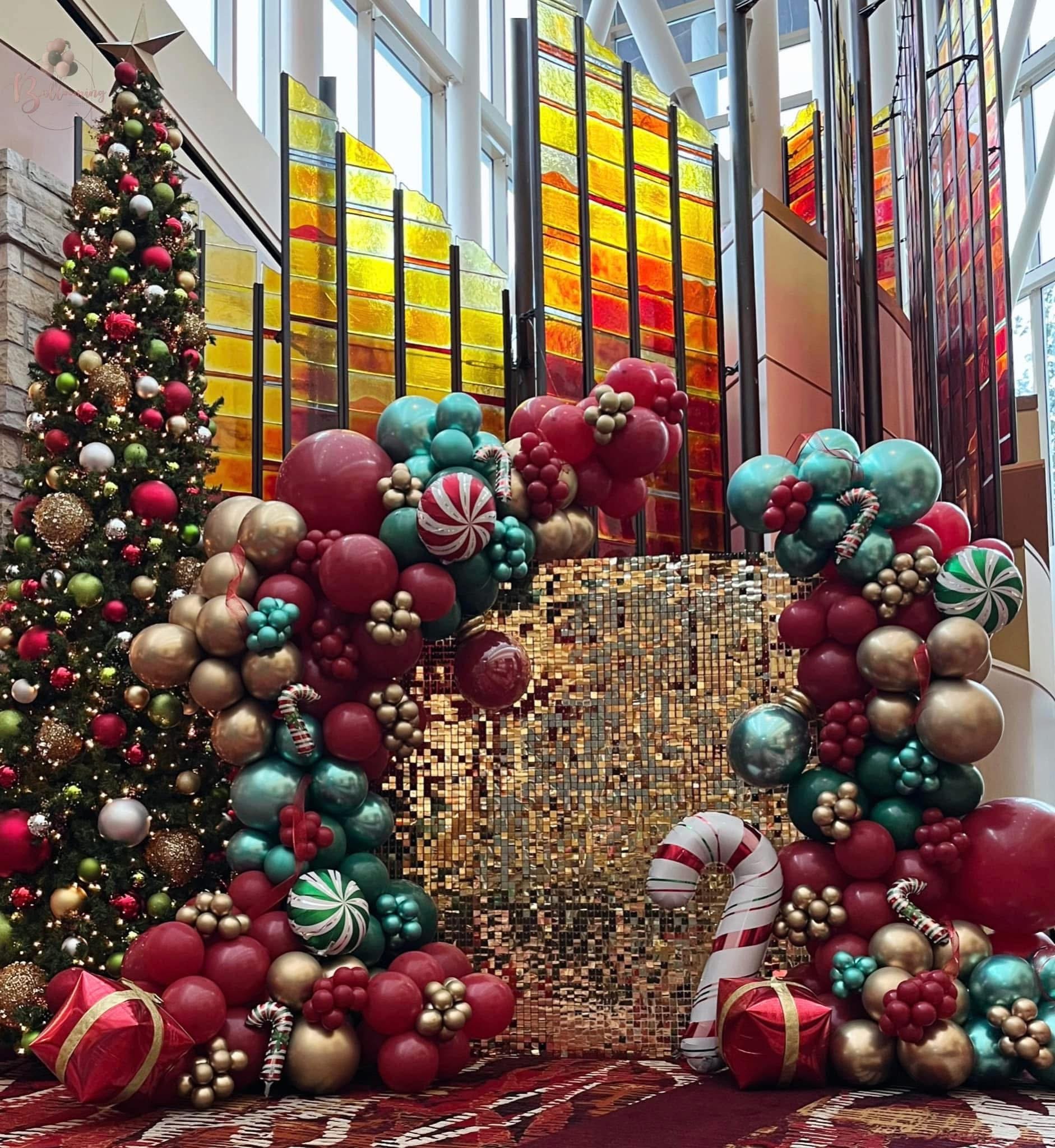 A Christmas scene featuring a decorated tree with ornaments and gold accents, holiday balloons in red, gold, and teal, candy cane props, a gold gift box, and a gold shimmering backdrop, all set inside a modern space with colorful stained glass window