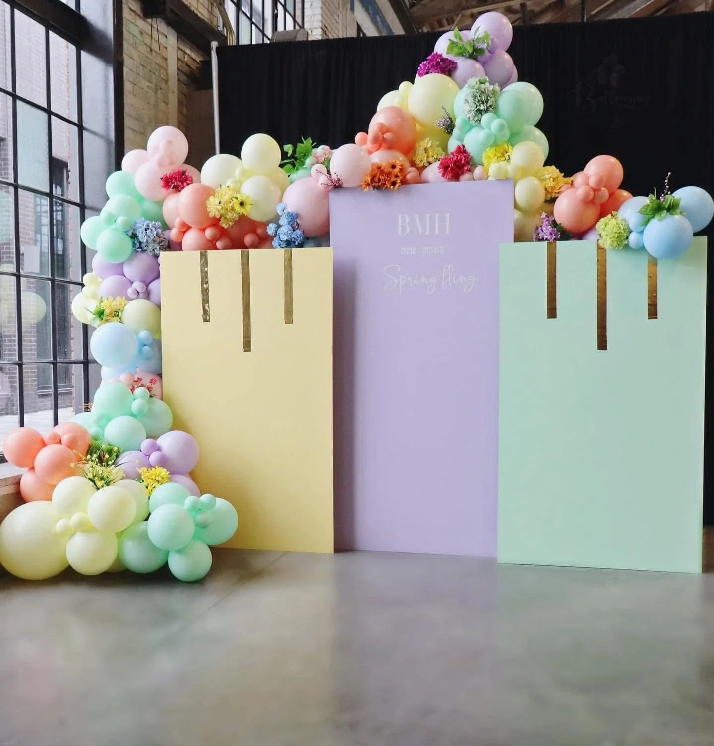 Colorful balloon arch with pastel-colored balloons and flowers, backdrop with a sign reading 'BMH presents Spring Fling' at an indoor event space.