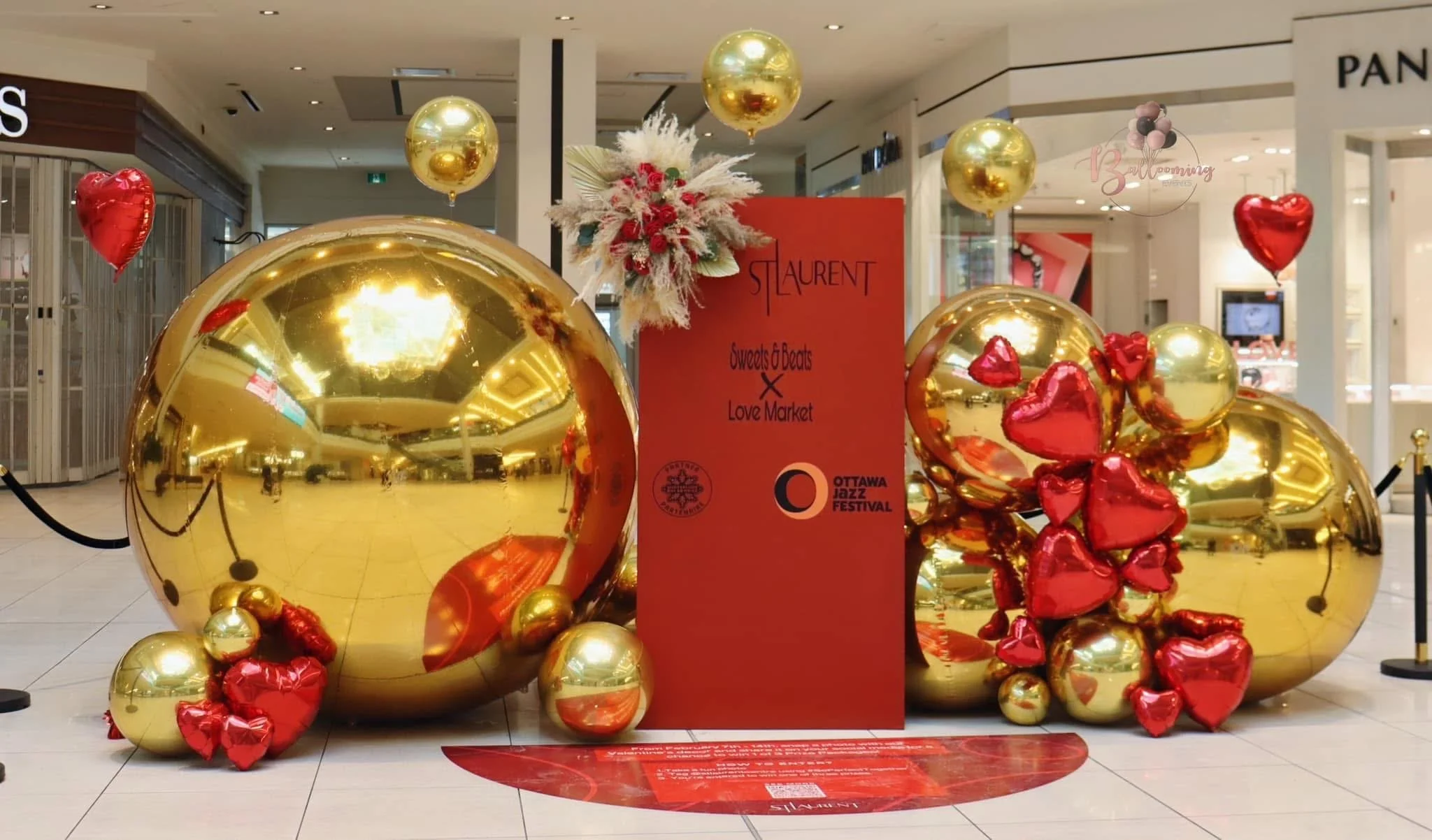 Decorative display with large gold and red balloons, a red sign with black text promoting a collaboration between Laurent Sweets & Beats and Love Market for Ottawa Jazz Festival, and a floral arrangement with white and red flowers in a shopping mall.
