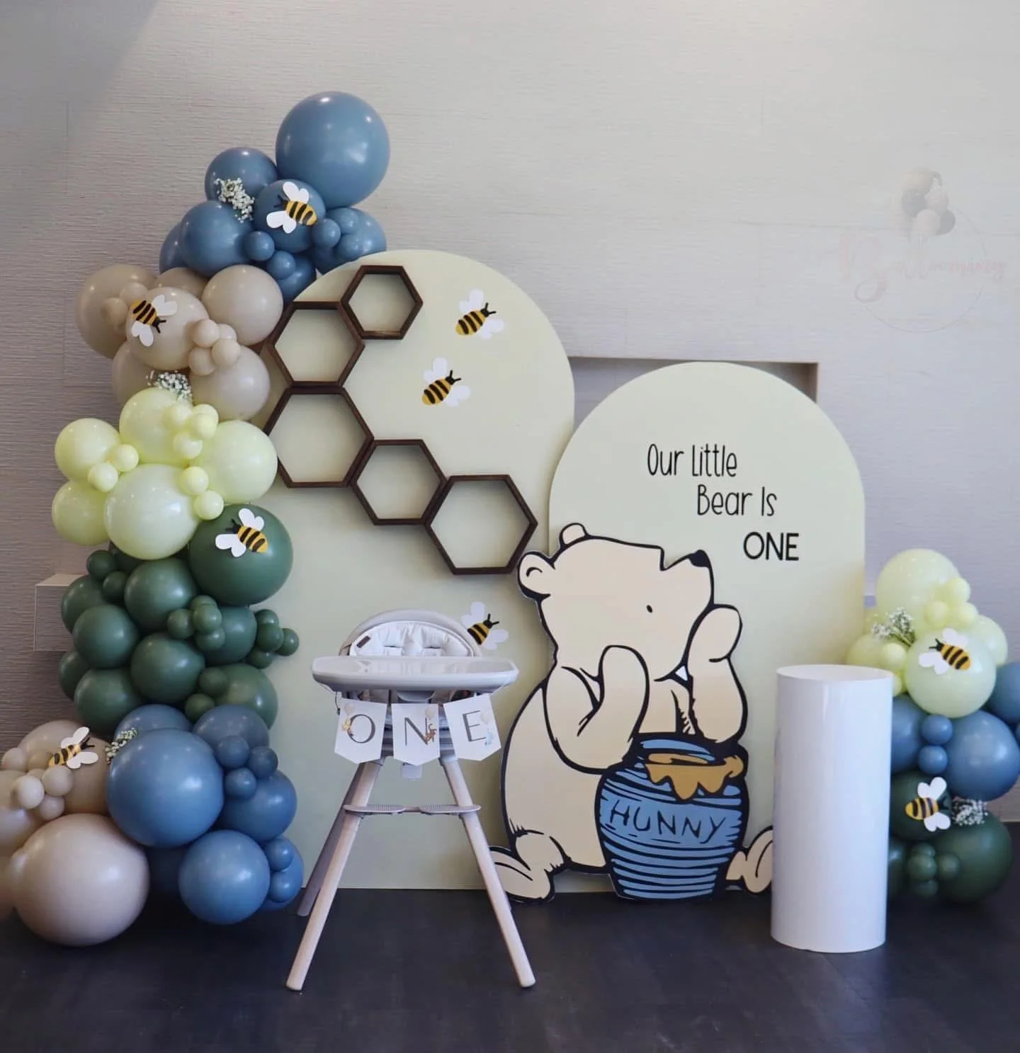 Decorative baby shower setup with balloon arrangements in green, blue, beige, and cream colors, painted bee stickers, honeycomb shapes, and a cartoon bear holding a honey jar with the text "Our Little Bear Is ONE." A white high chair with a "ONE" ban