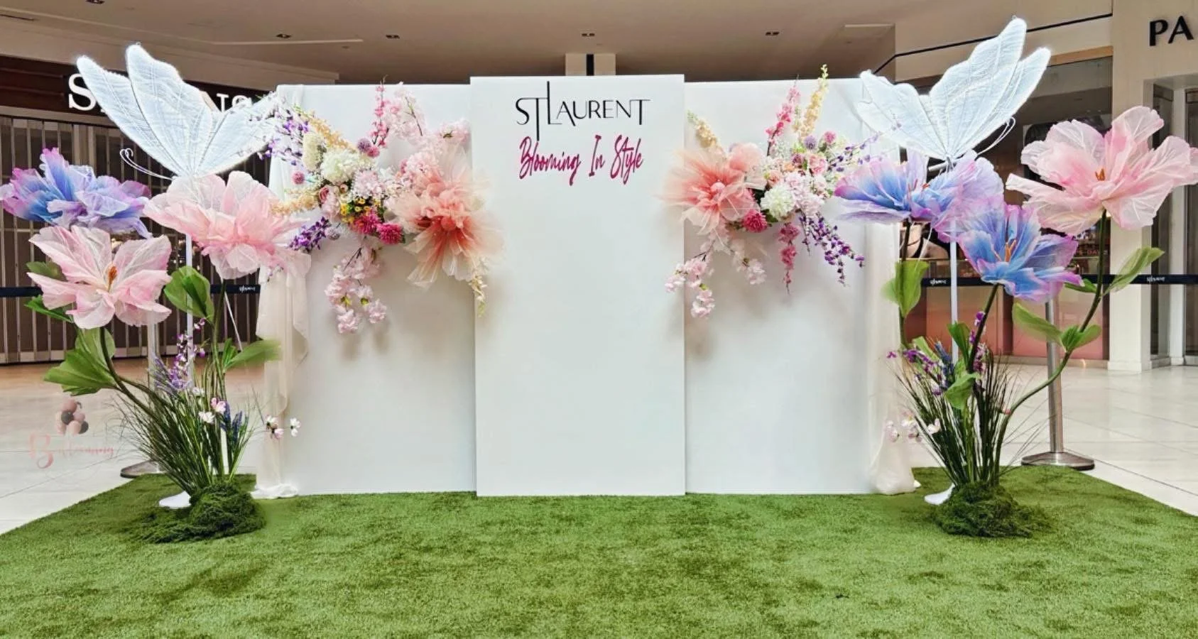 Decorative floral display with pink and purple flowers, white butterflies, and a white backdrop with the words 'ST LAURENT Booming In Style' at a shopping mall.
