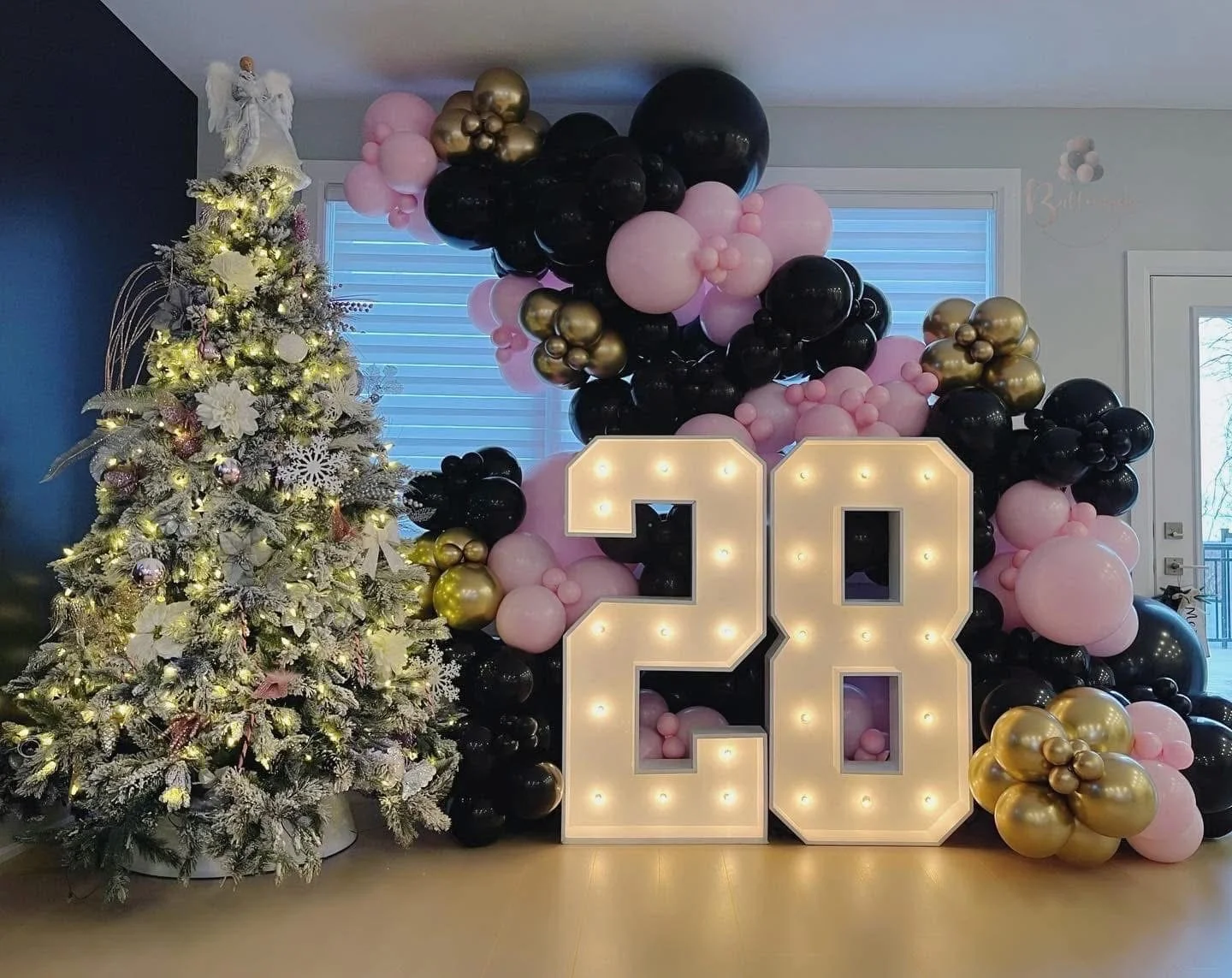 Decorative setup with a white Christmas tree adorned with lights and white ornaments on the left, and a balloon arrangement with pink, black, and gold balloons behind large illuminated number '20' for a celebration, possibly a 20th birthday or annive