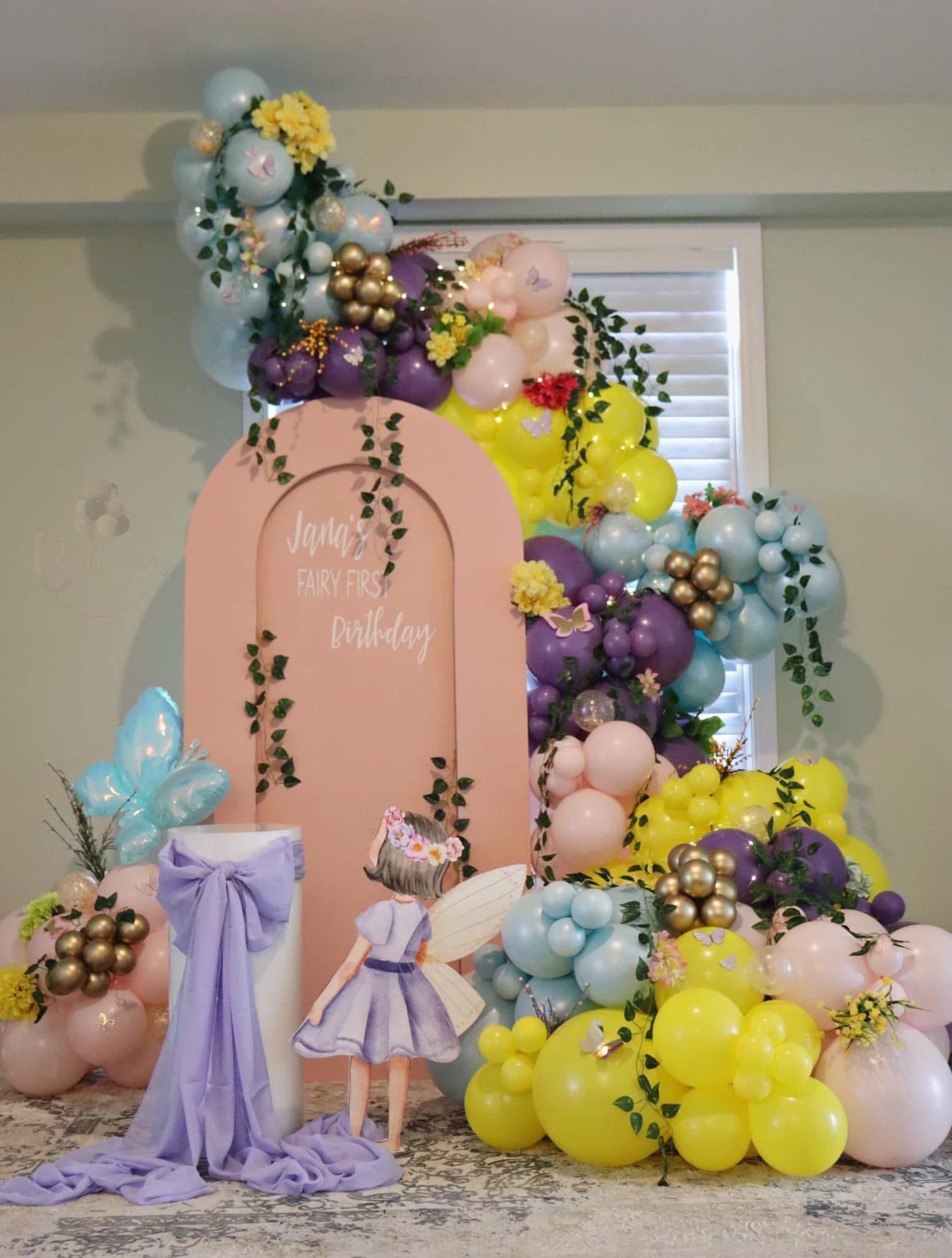 Decorations for a child's first birthday party with pastel-colored balloons, flowers, greenery, and a fairy-themed backdrop featuring a girl with fairy wings and a floral headband.