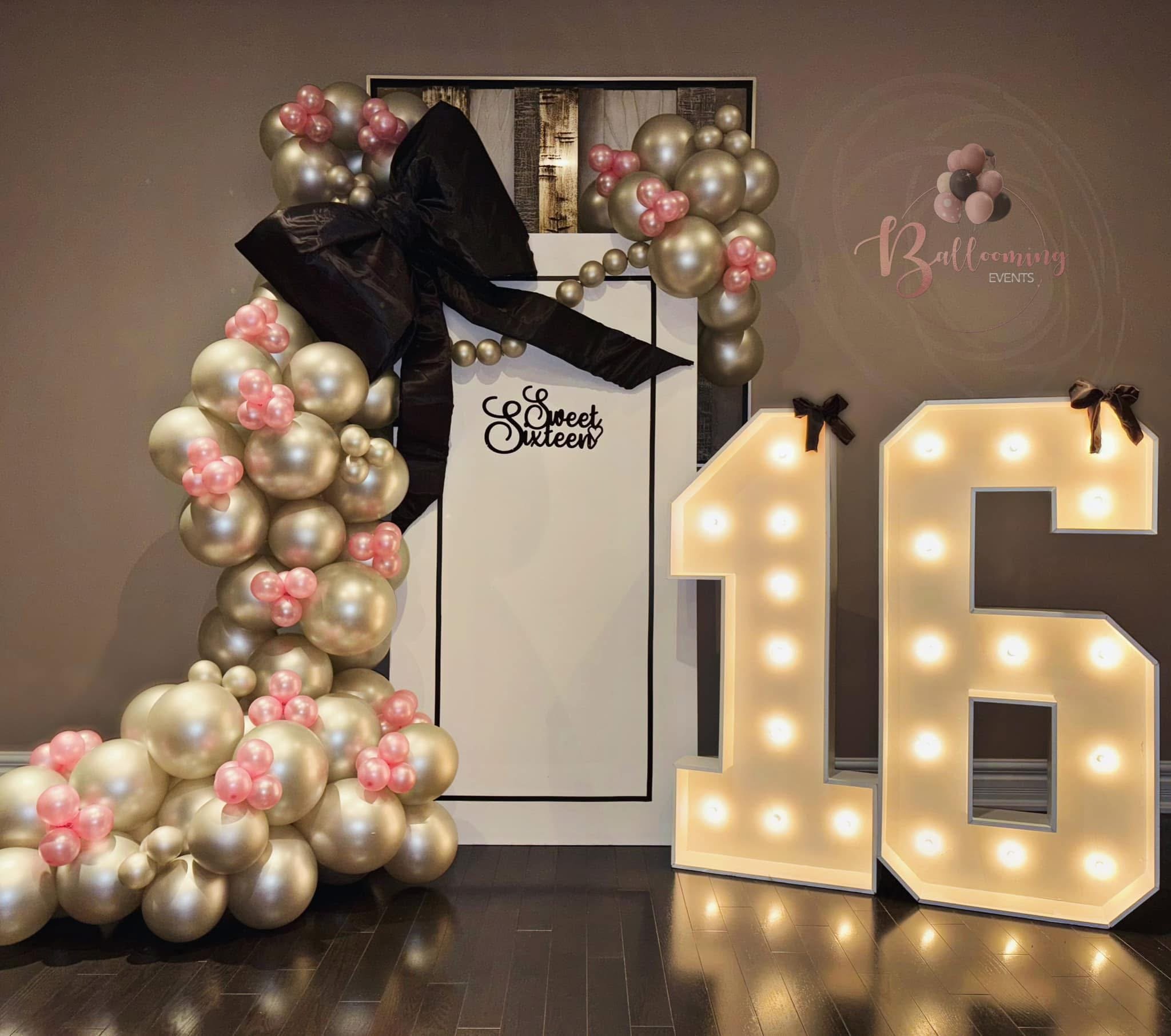 Decorations for an 16th birthday celebration featuring a large arrangement of gold and pink balloons, a black ribbon bow, a white door with a sign that says 'Sweet Sixteen,' illuminated marquee numbers '16' with black bows, and a wall sign indicating