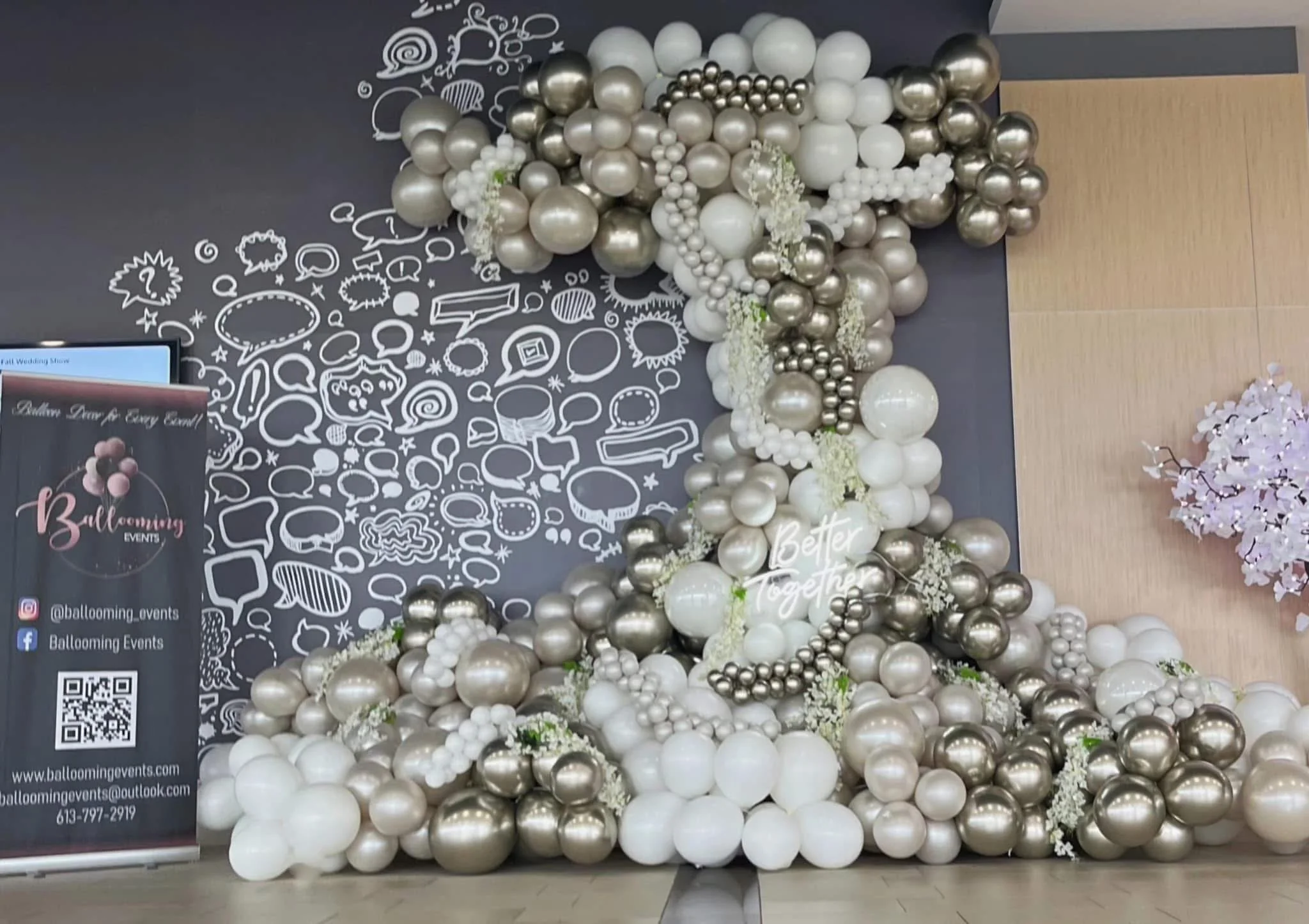 Decorative arrangement of white, silver, and pearl-colored balloons with a neon sign that says 'Better Together' against a black wall with white doodles, and a pink floral display to the right.