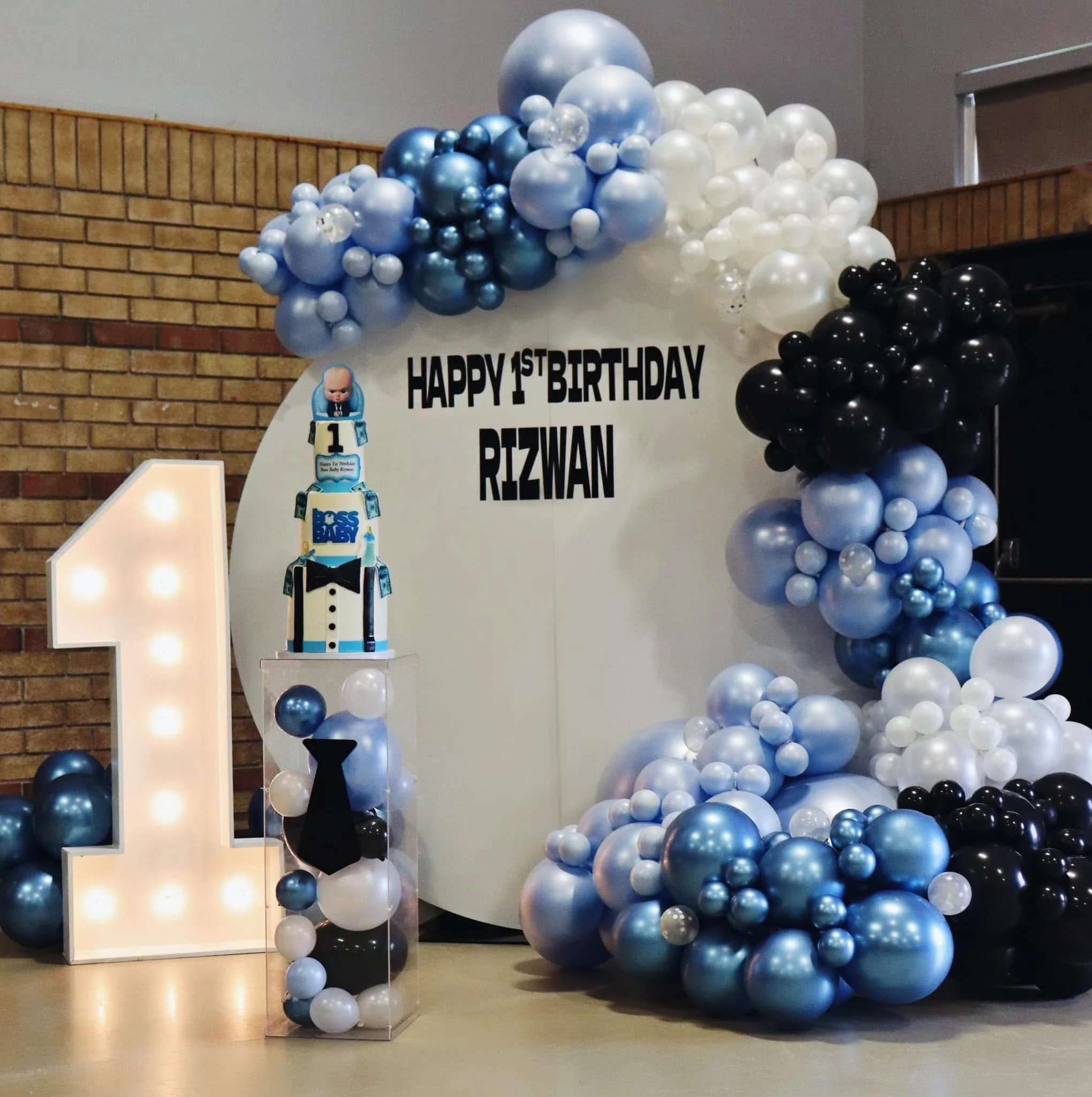 First birthday celebration decor for Rizwan with a large light-up number one, black, white, blue, and pearl balloons, a birthday cake with a baby figure on top, and a sign saying "Happy 1st Birthday Rizwan".