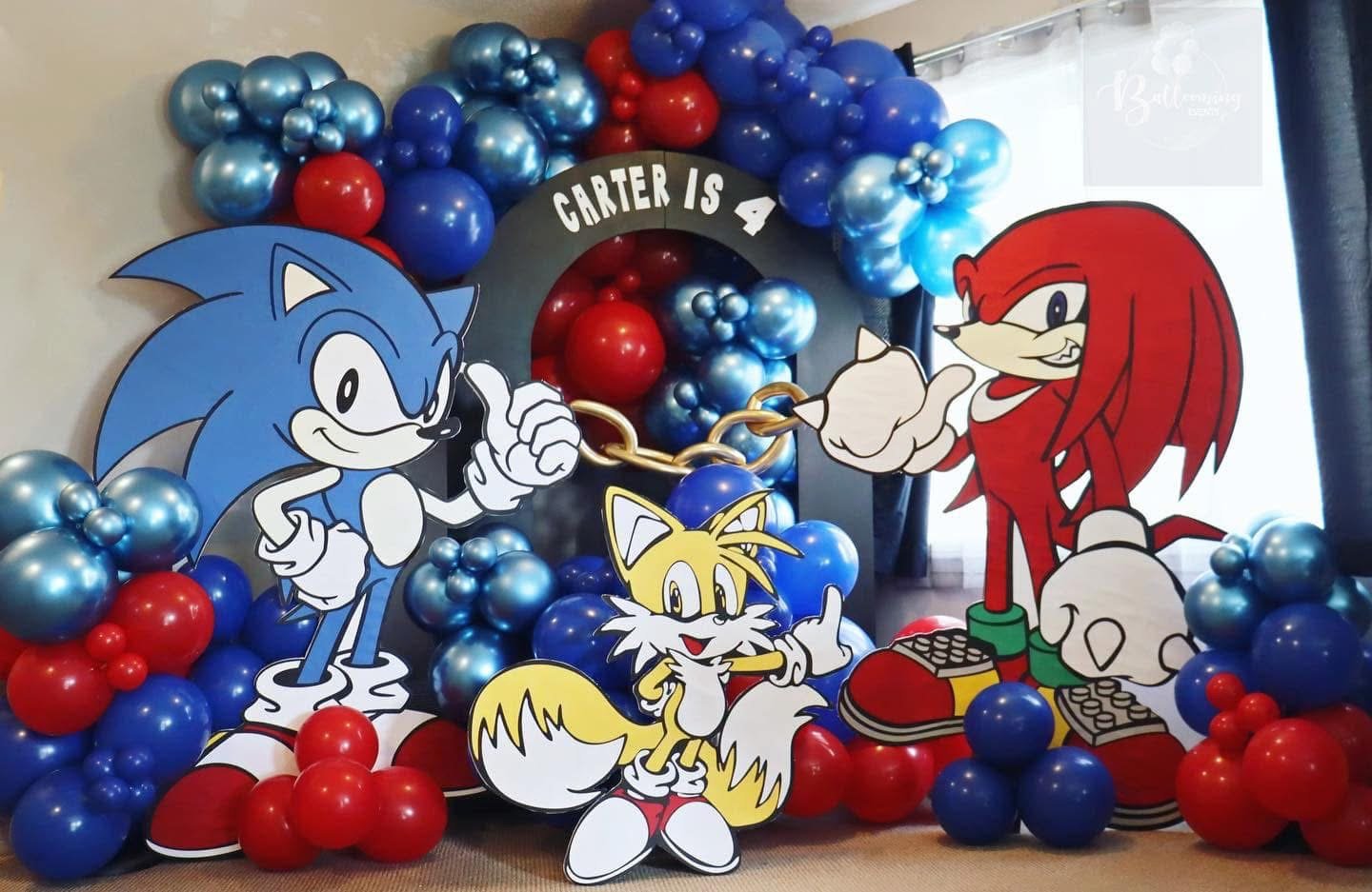 Decorations for a Sonic the Hedgehog themed birthday party, featuring large cutouts of Sonic, Tails, and Knuckles, surrounded by blue and red balloons, with a black arch that reads "Carter is 4" and a golden chain connecting Sonic and Knuckles.