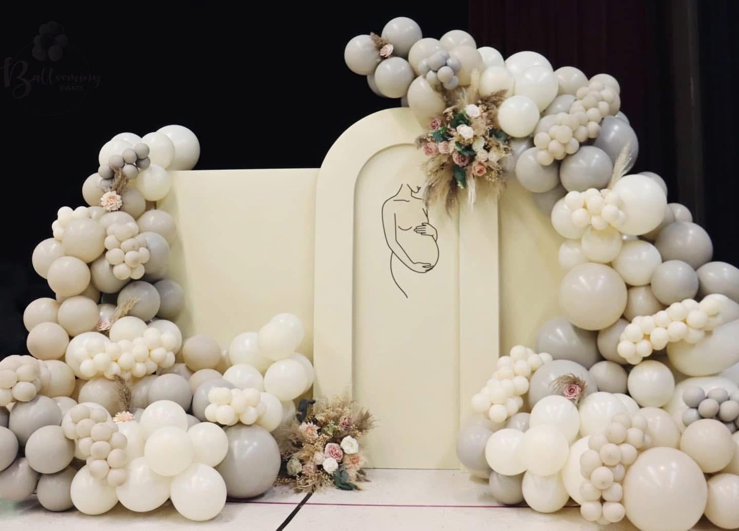 Decorative backdrop with cream-colored balloons arranged around a simple outline drawing of a pregnant woman, accented with pink and white flowers and dried foliage.