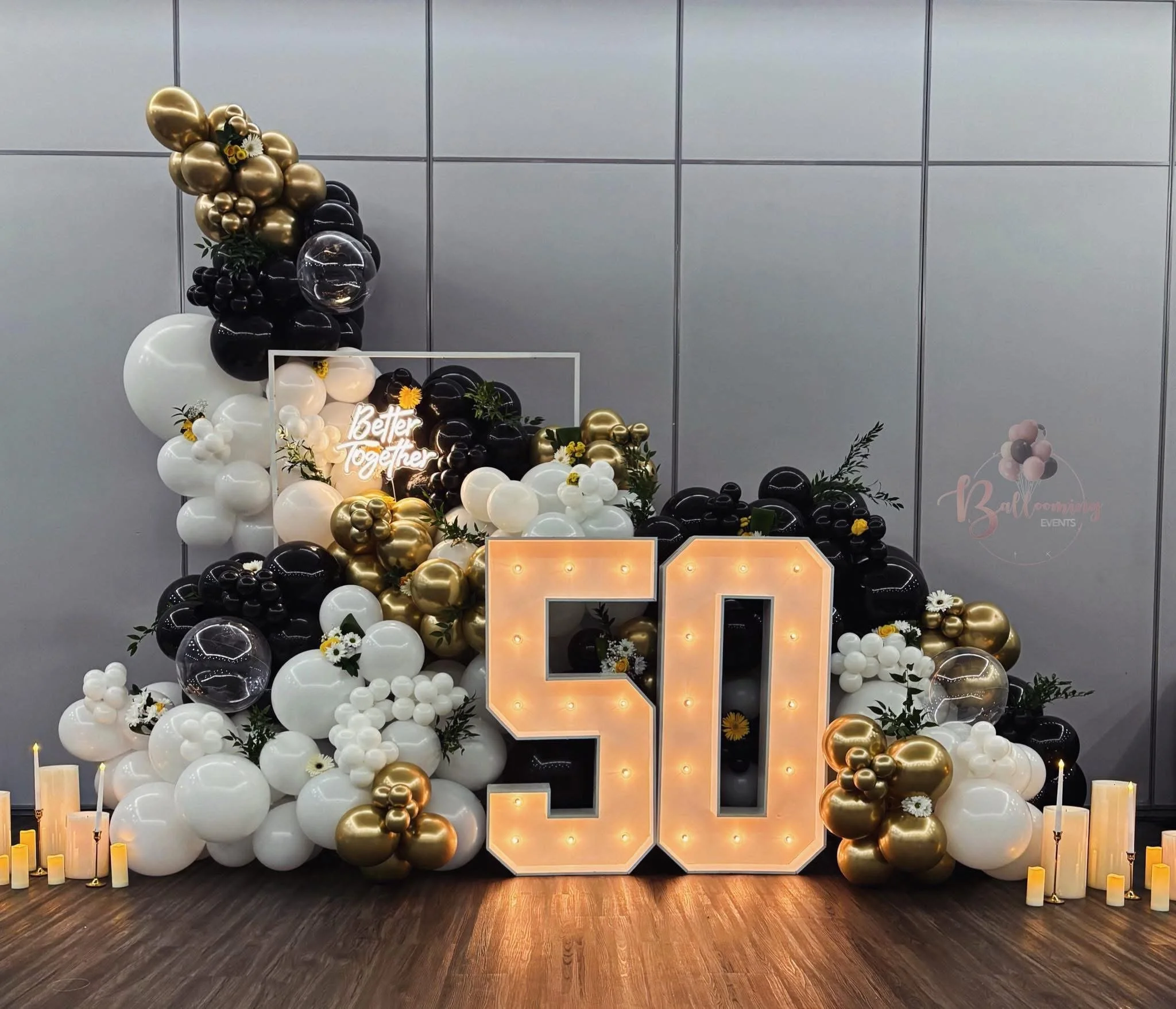 A celebration decoration for a 50th birthday with large illuminated number caulk, black, white, and gold balloons, candles, and greenery on a wooden floor against a gray wall.