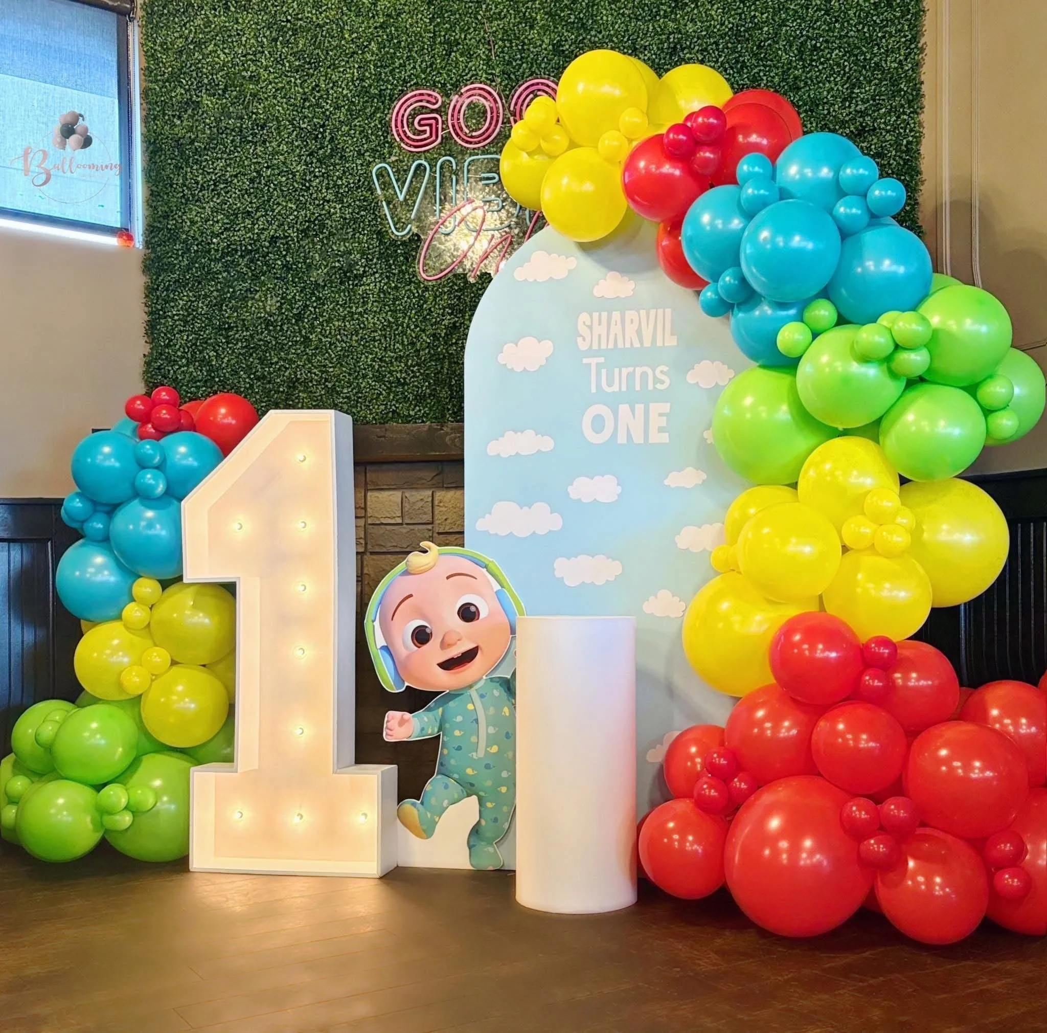 Decorative birthday display with a large illuminated number one, colorful balloon arrangements in red, yellow, green, and blue, a cartoon character cutout, and a backdrop that reads "Sharvil Turns One" with clouds, against a green leafy wall.