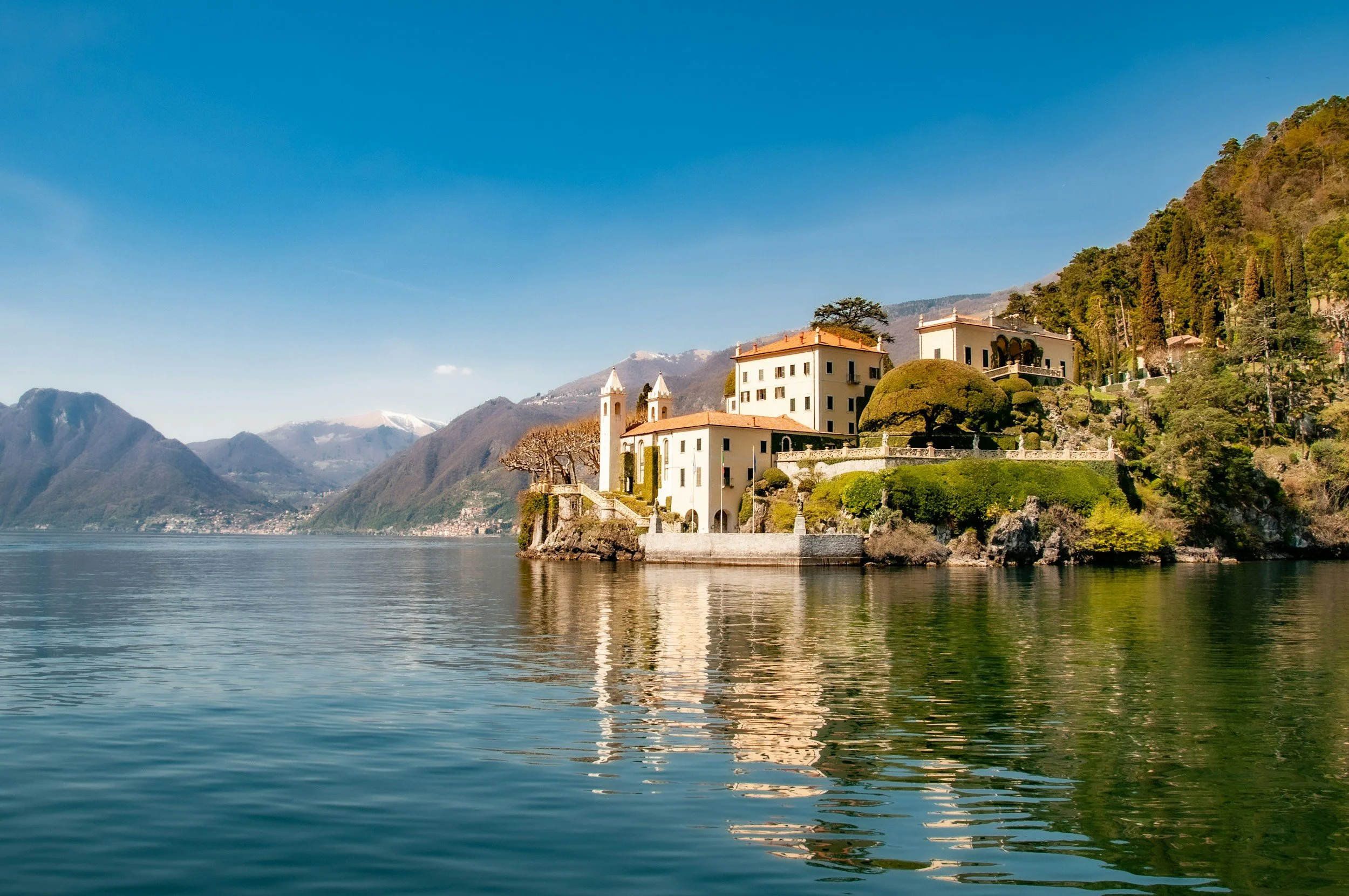 A scenic view of a lakeside villa with green lawns and trees, set against distant mountains under a clear blue sky.