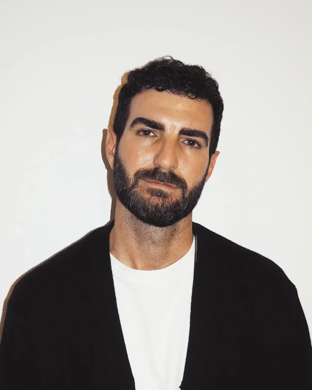 Portrait of a man with dark, curly hair and a beard, wearing a black cardigan over a white t-shirt, standing against a plain white background.
