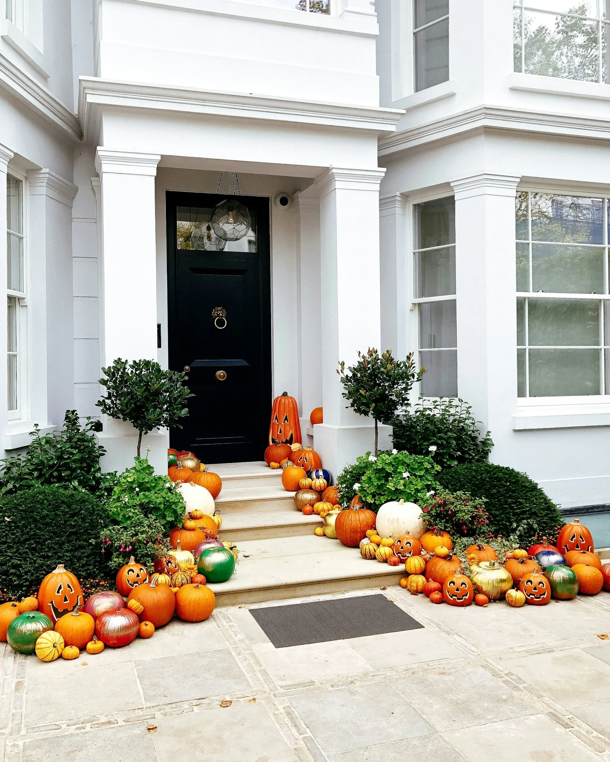 Front porch decorated for Halloween with pumpkins and gourds of various sizes and colors, including traditional orange, white, green, and gold. Some pumpkins are carved with jack-o'-lantern faces. The porch has white steps and a black door with a door knocker, and there are two small bushes on either side of the steps.