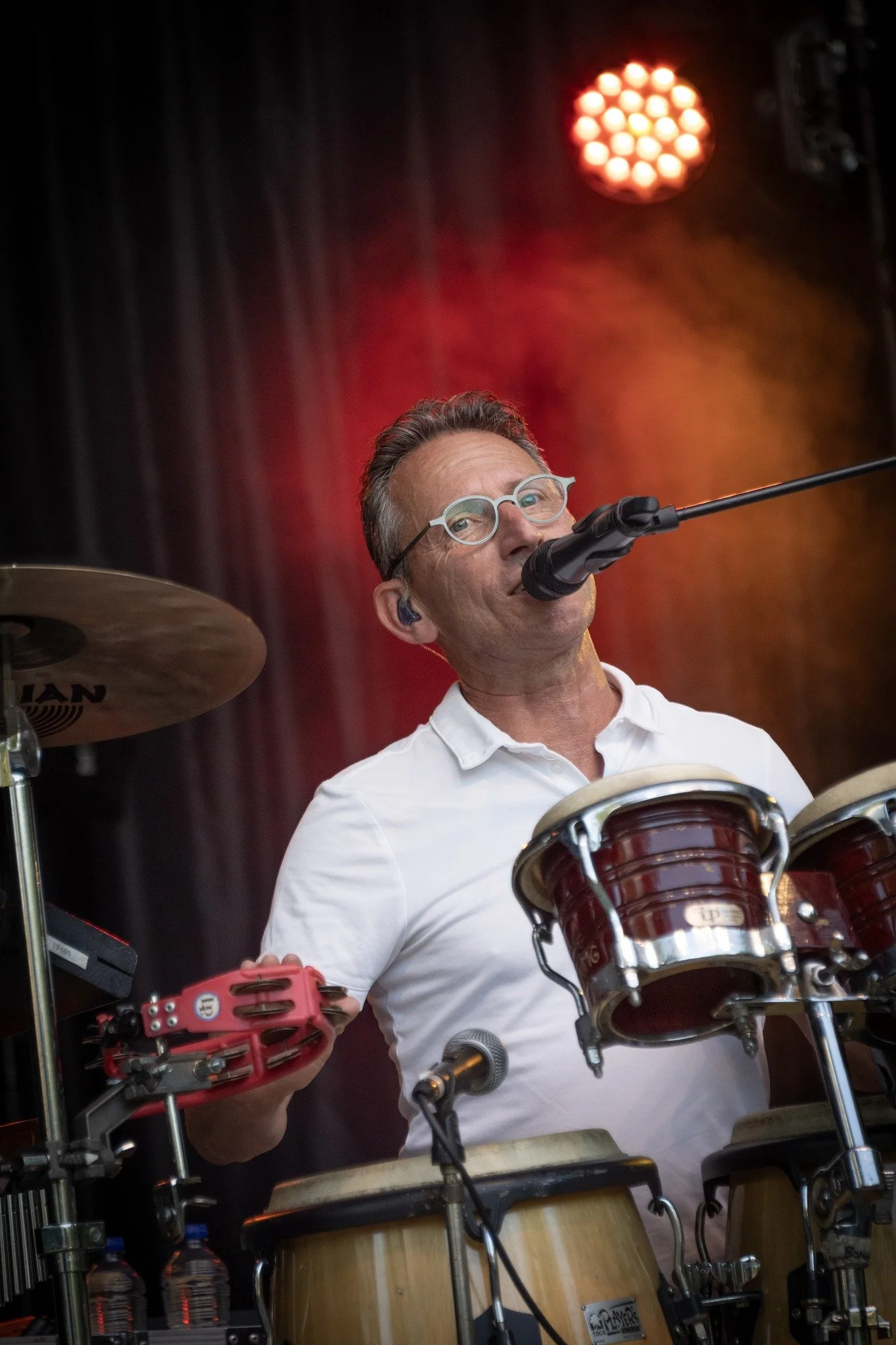 Peter van Whenever zingt live in een microfoon terwijl hij percussie speelt, met bril en wit poloshirt, op het podium.