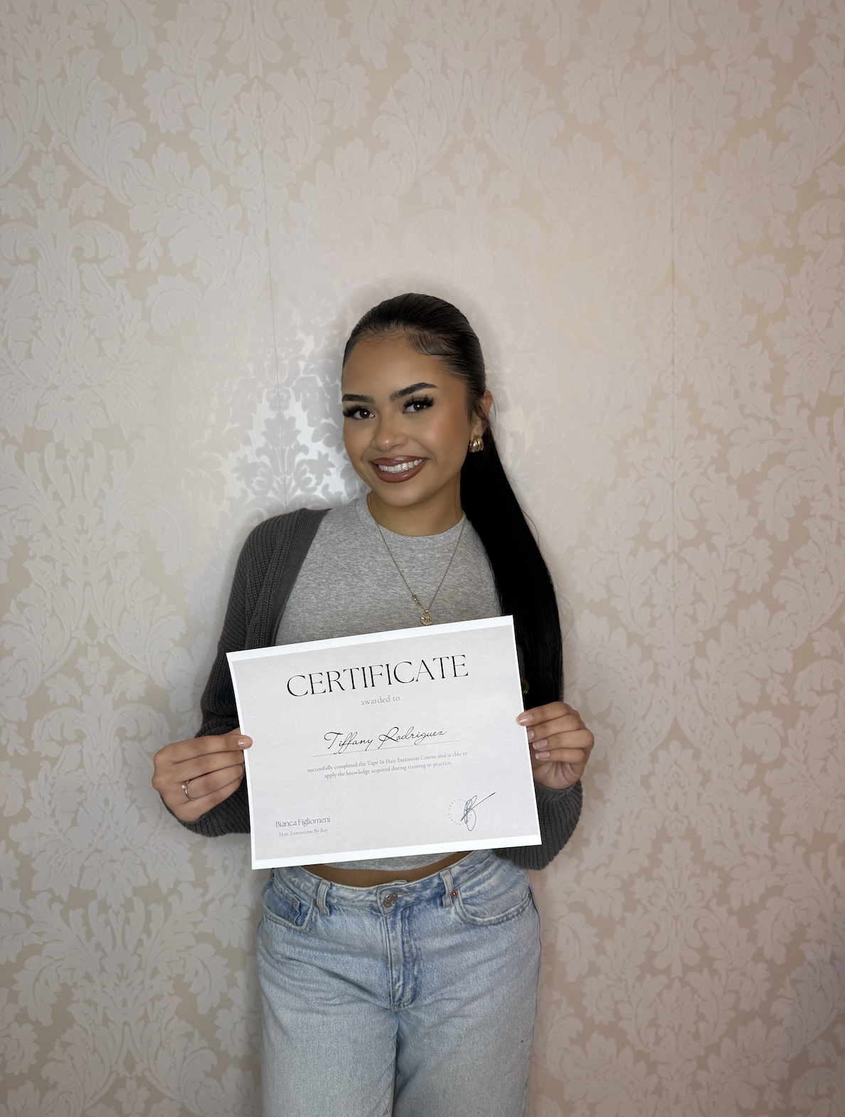 Young woman with dark hair, smiling, holding a certificate, standing against a cream floral wallpaper background.