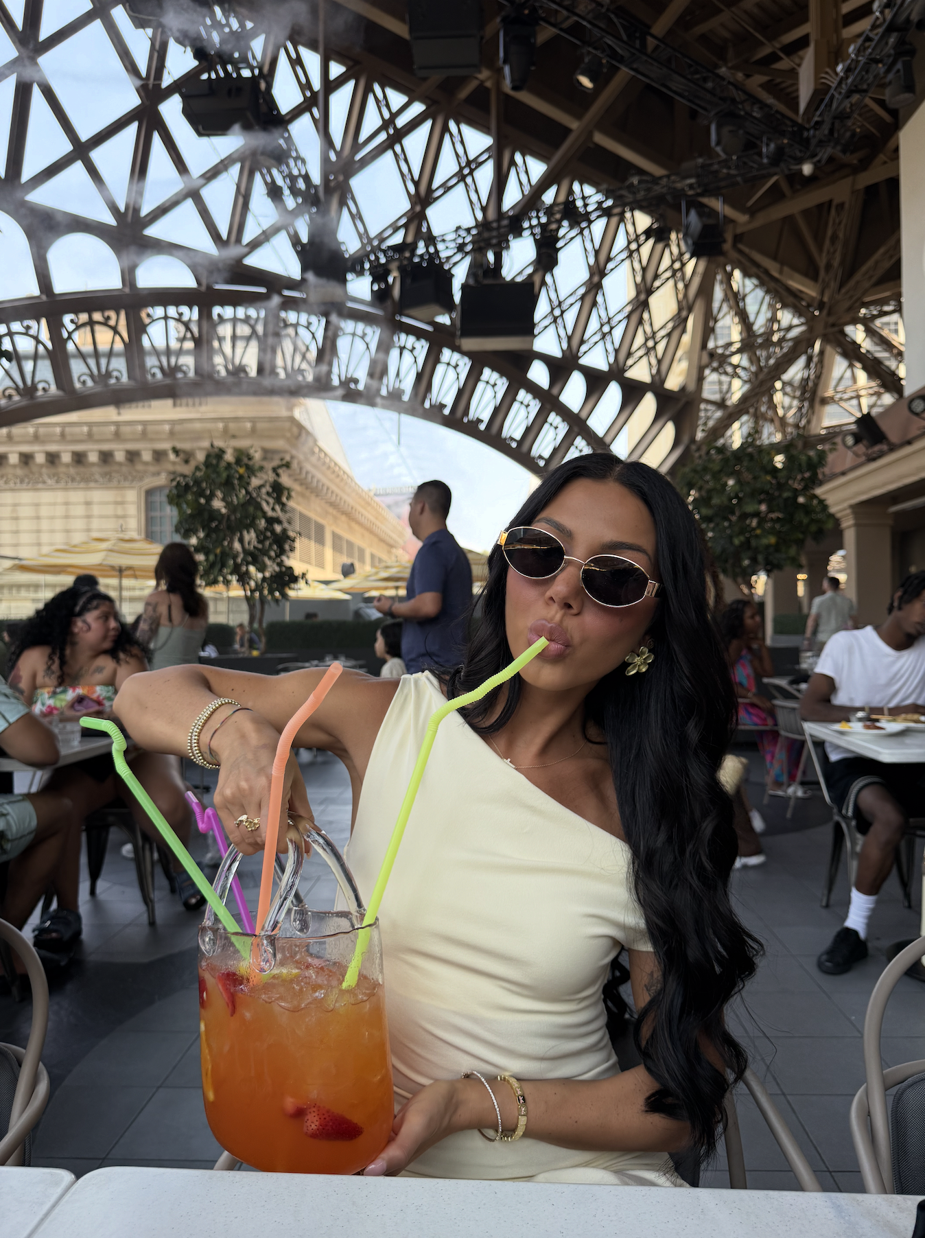A woman with long dark hair, wearing sunglasses and a light yellow dress, is sitting at an outdoor restaurant with a large drink and colorful straws, making a playful pouty face.