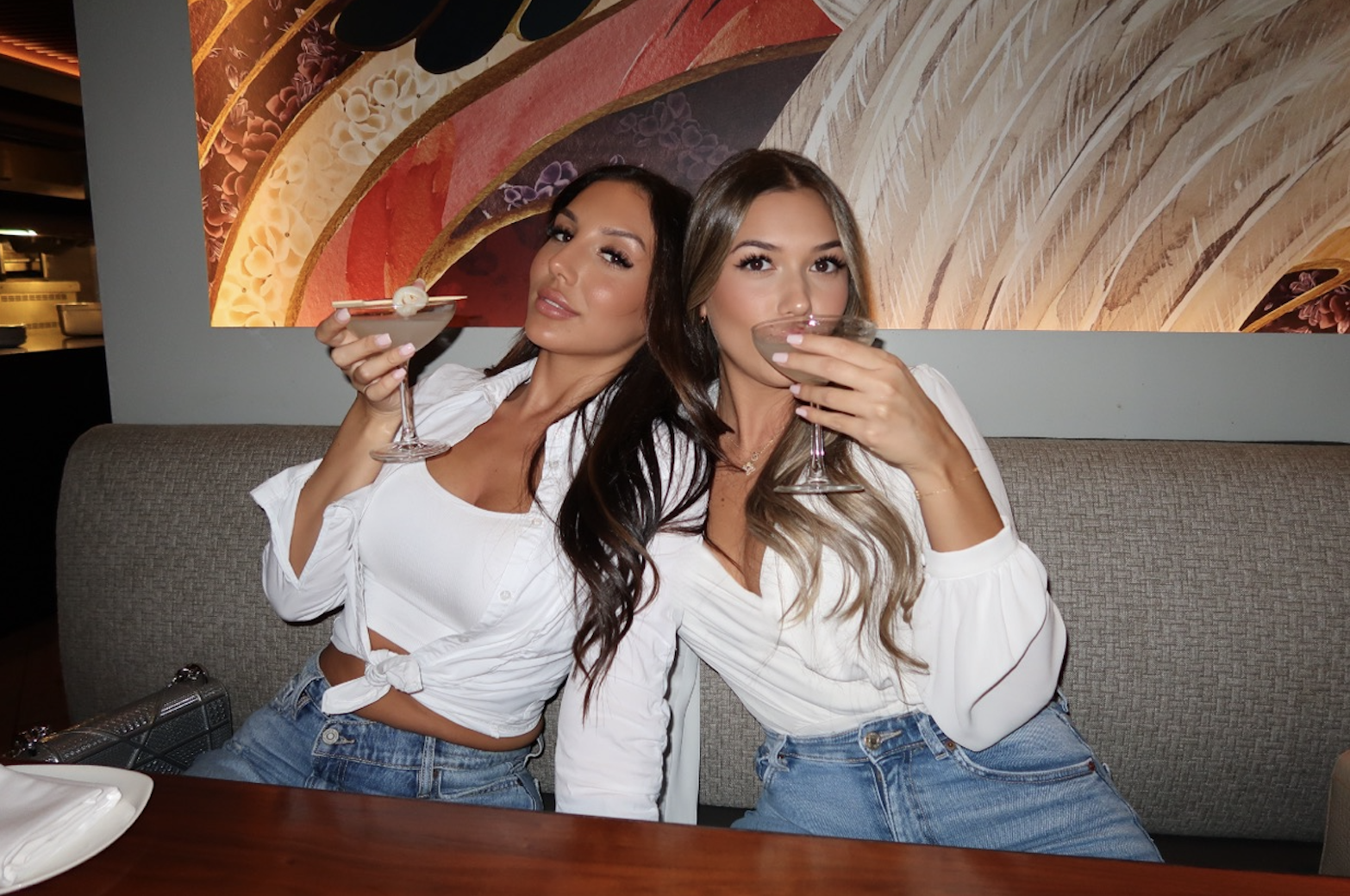 Two women sitting on a restaurant booth, holding cocktail glasses and posing for the camera. One woman has dark hair, the other has blonde hair. They are dressed in white tops and blue jeans. There is a colorful abstract wall art behind them.