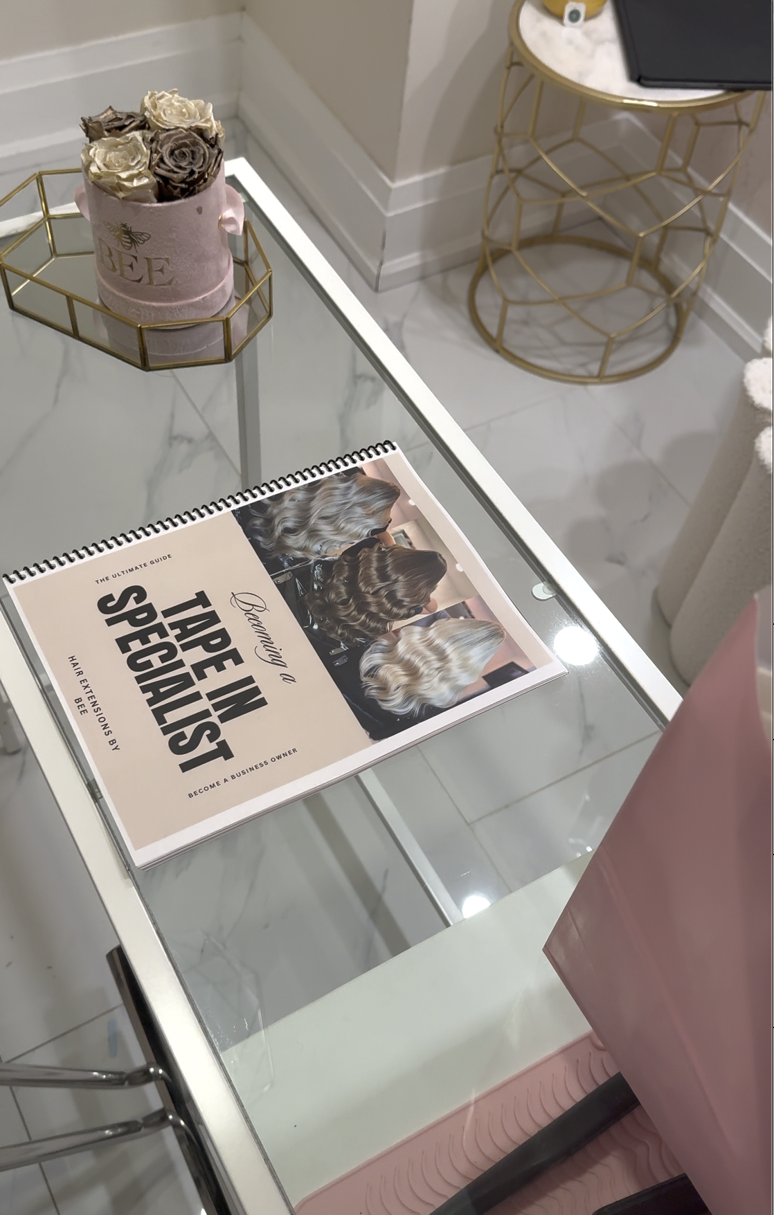 A glass-topped table with a pamphlet titled 'Becoming a Tape in a Specialist' showing images of different hairstyles, and a pink flower arrangement in a pink container with a bee illustration, in a room with white walls and gold-colored furniture.
