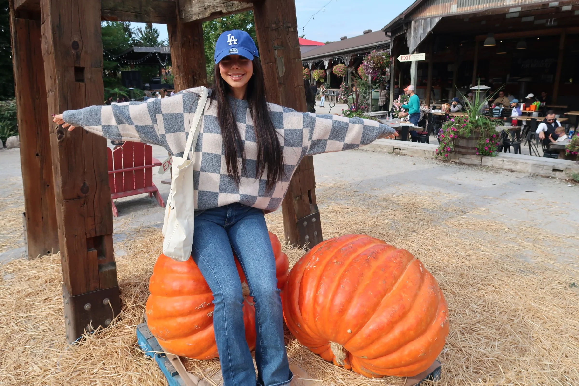 A young woman with long dark hair, wearing a blue Los Angeles Dodgers cap, grey and white checkered sweater, and jeans, sits on a large orange pumpkin with her arms outstretched in a welcoming pose at a pumpkin patch. In the background, there are people dining and walking outside a rustic restaurant or café with hanging flowers and outdoor seating.