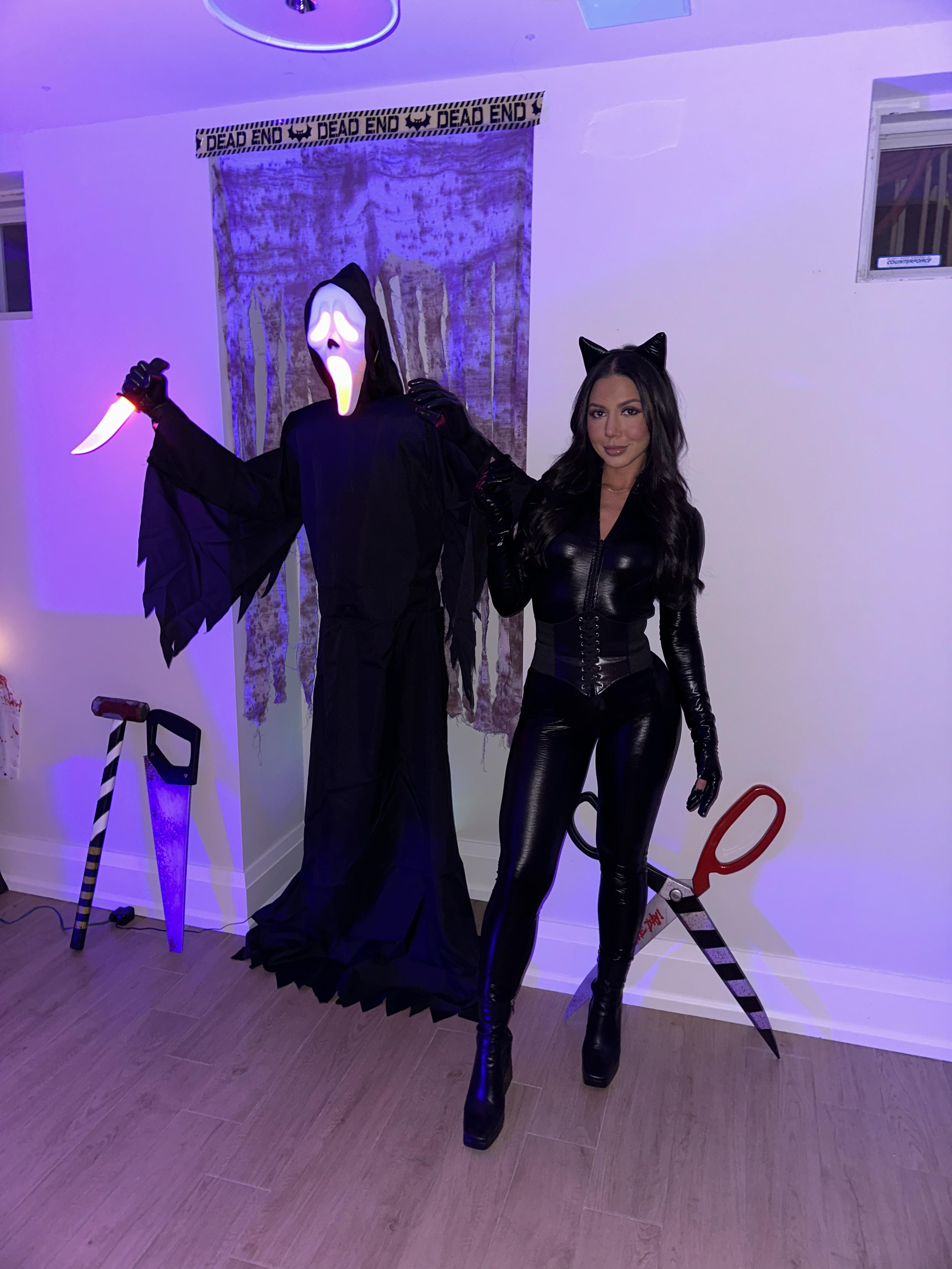 Woman dressed in all black leather cat costume with cat ears hat, standing next to a hanging spooky ghost or Grim Reaper figure with a white skull mask and glowing mouth, holding a prop knife. Scissors and a bat-shaped object are on the floor nearby.