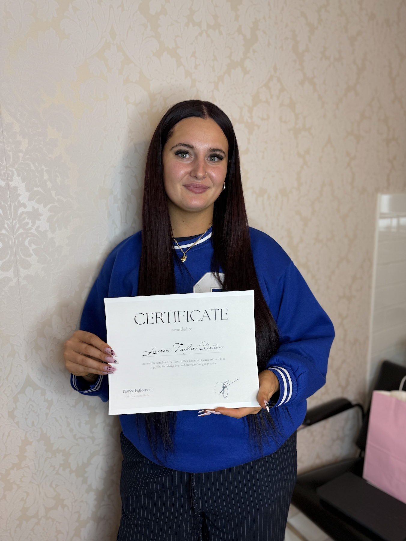 A woman with long dark hair, wearing a blue sweatshirt, is smiling while holding a certificate in front of her. She has a light skin tone and is standing against a cream-colored wall with a floral pattern.